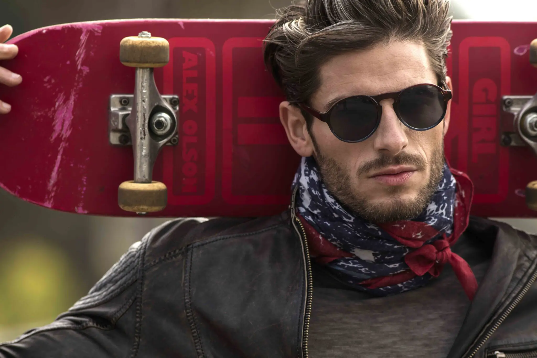 Man wearing sunglasses and a leather jacket holding a red skateboard behind his head.