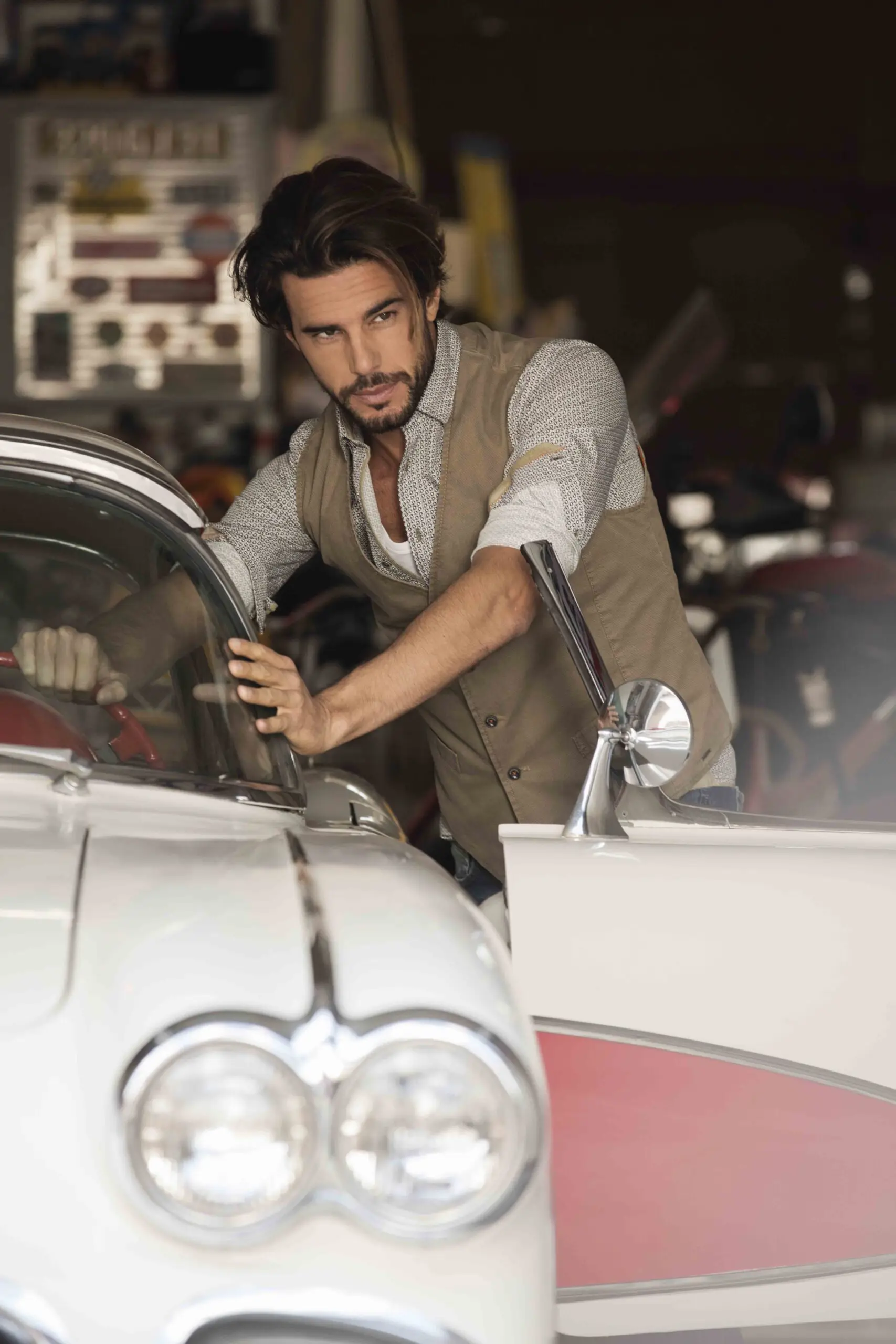 Man with dark hair and beard wearing a beige vest leaning on the open door of a classic white car.