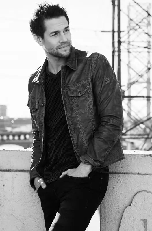 Man with short dark hair wearing a casual jacket and black shirt, leaning against a concrete railing outdoors.