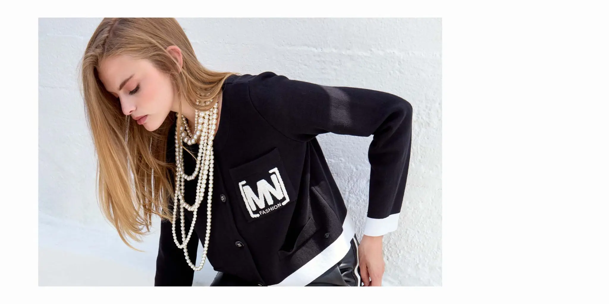 Woman with long blonde hair wearing a black cardigan with white cuffs and a pearl necklace, posing against a white textured wall.
