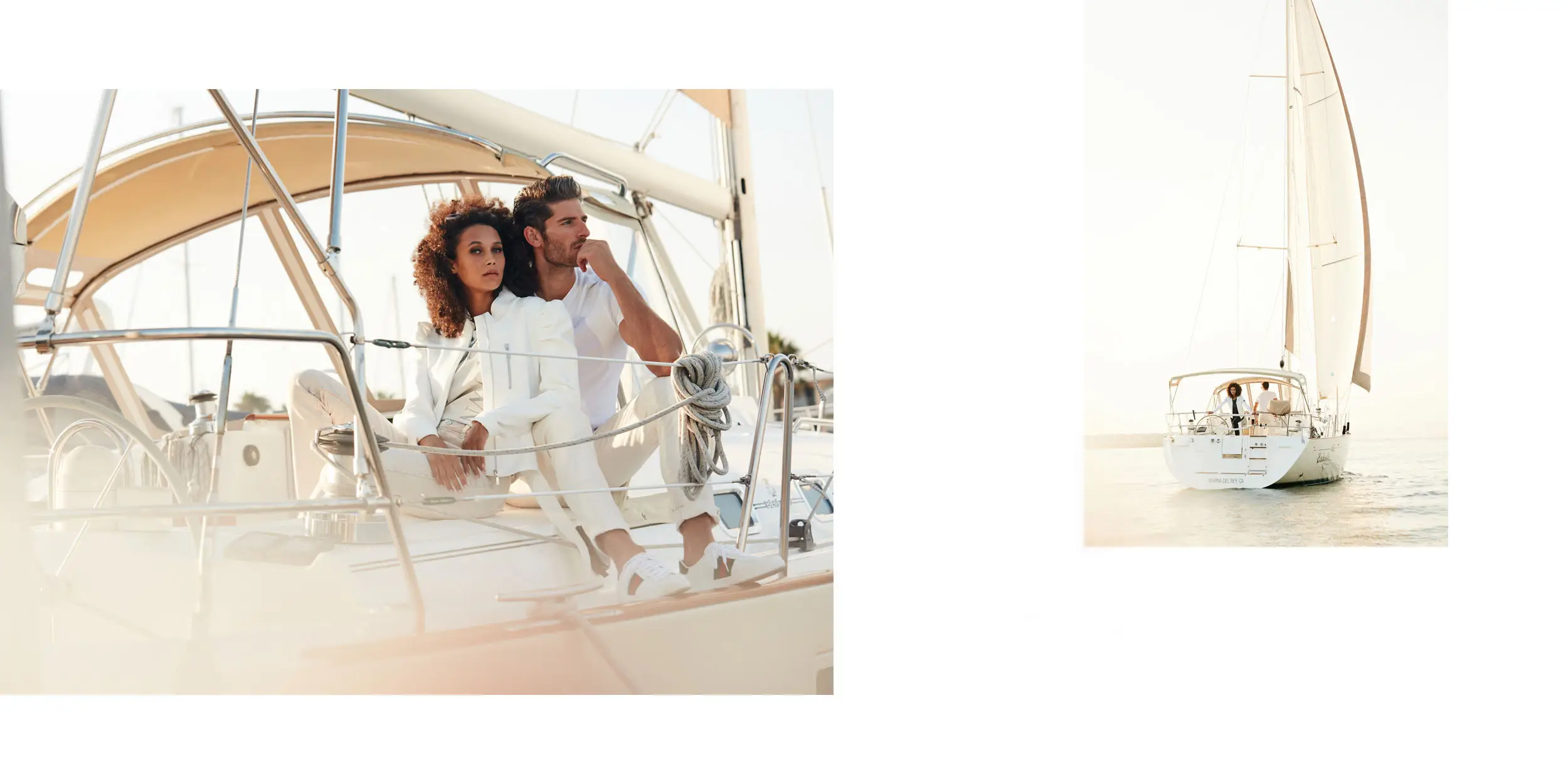 Couple dressed in white sitting on the deck of a sailboat with sails raised on calm water.