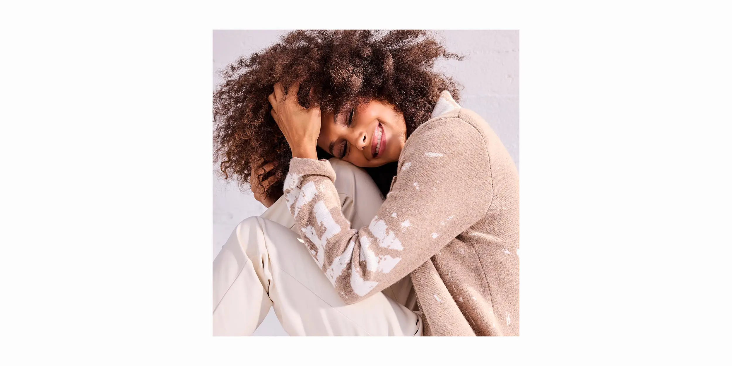 Smiling woman with curly hair resting her head on her knees, wearing a beige sweater and light pants.