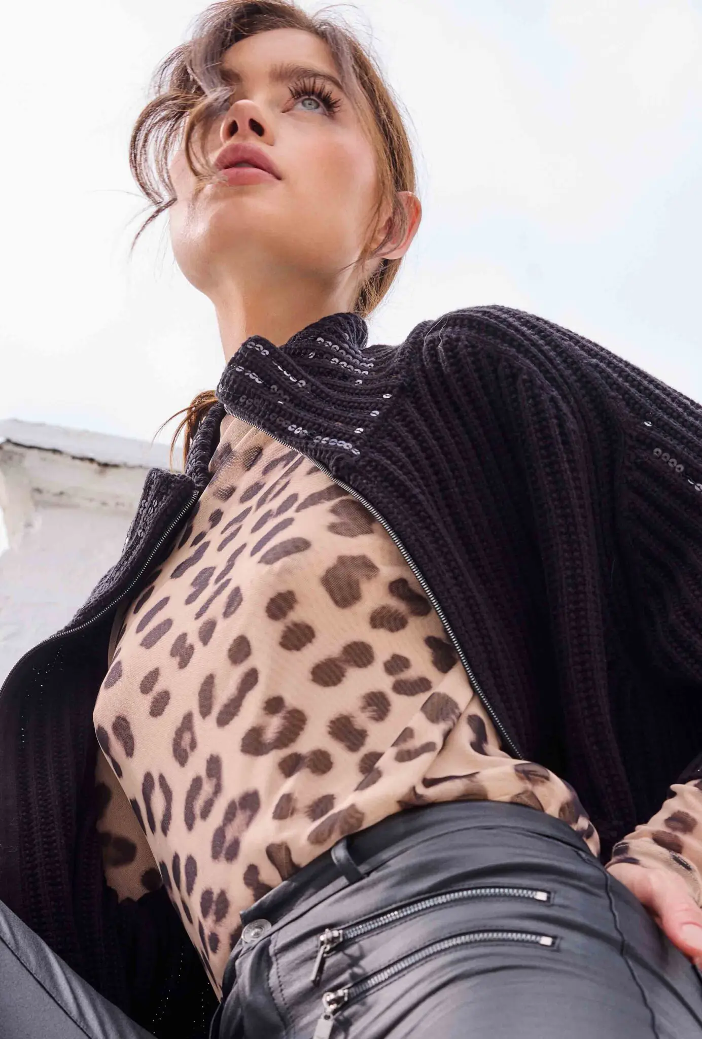 Low-angle view of a woman wearing a leopard print top, black knit cardigan, and black leather pants with zipper details.