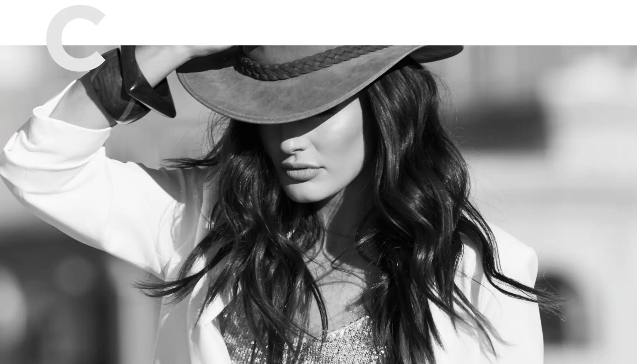 Woman with long wavy hair wearing a fedora hat tilted over her eyes, a white blazer, and a sequined top.