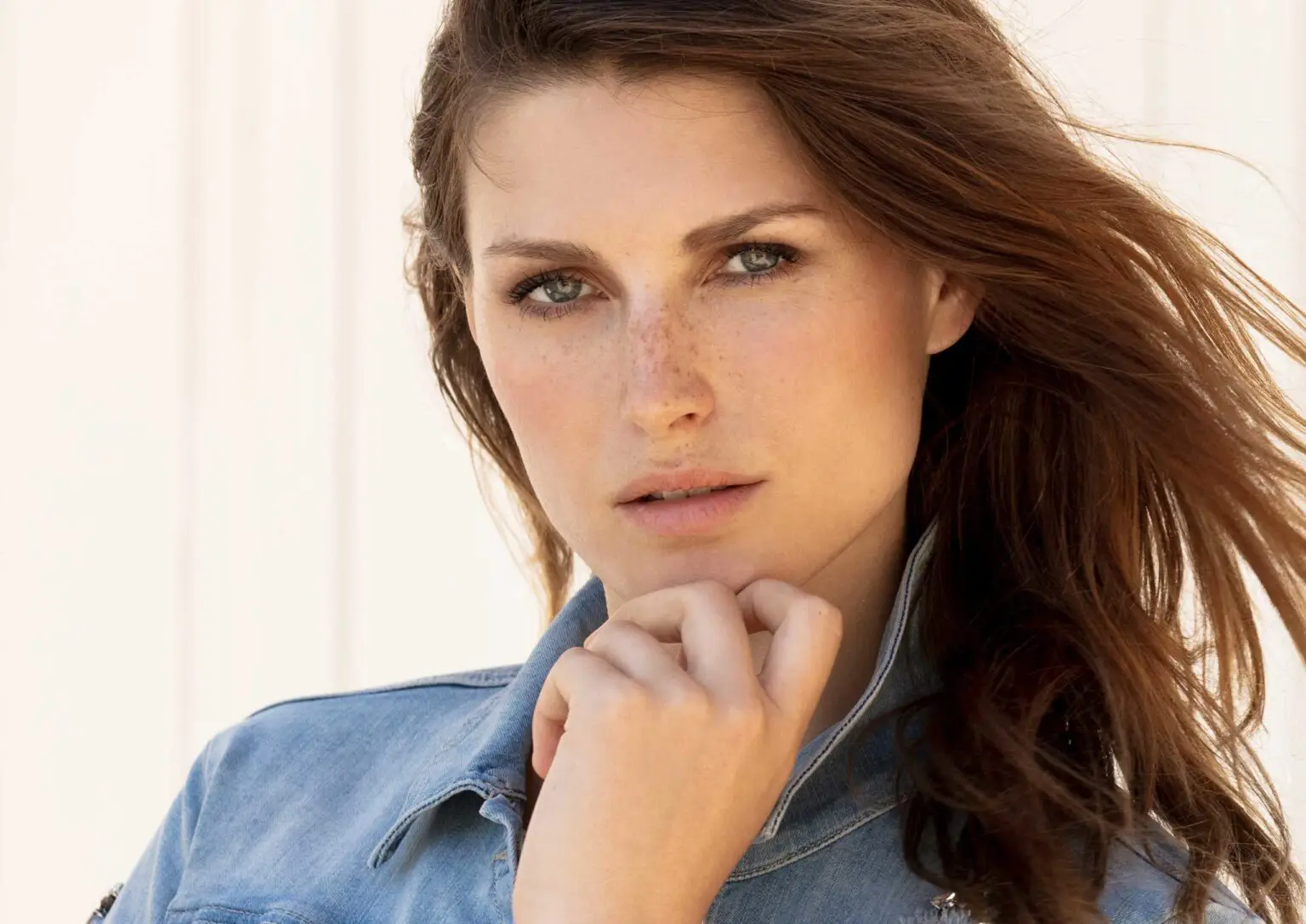 Close-up of a woman with freckles and windblown brown hair wearing a light denim jacket, resting her chin on her hand.