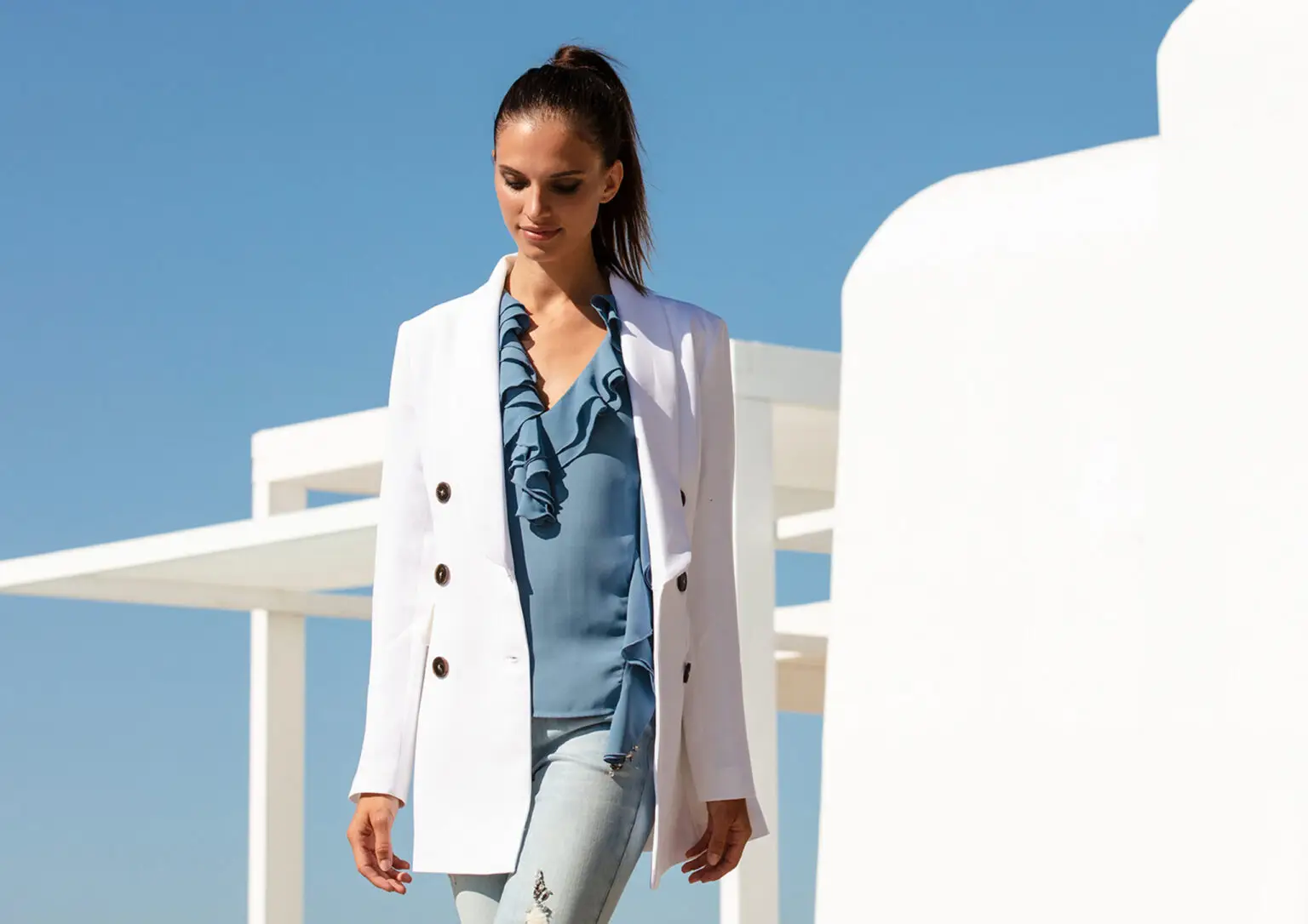 Woman wearing a white blazer over a blue ruffled blouse and light blue jeans standing outdoors against a clear blue sky and white modern architecture.