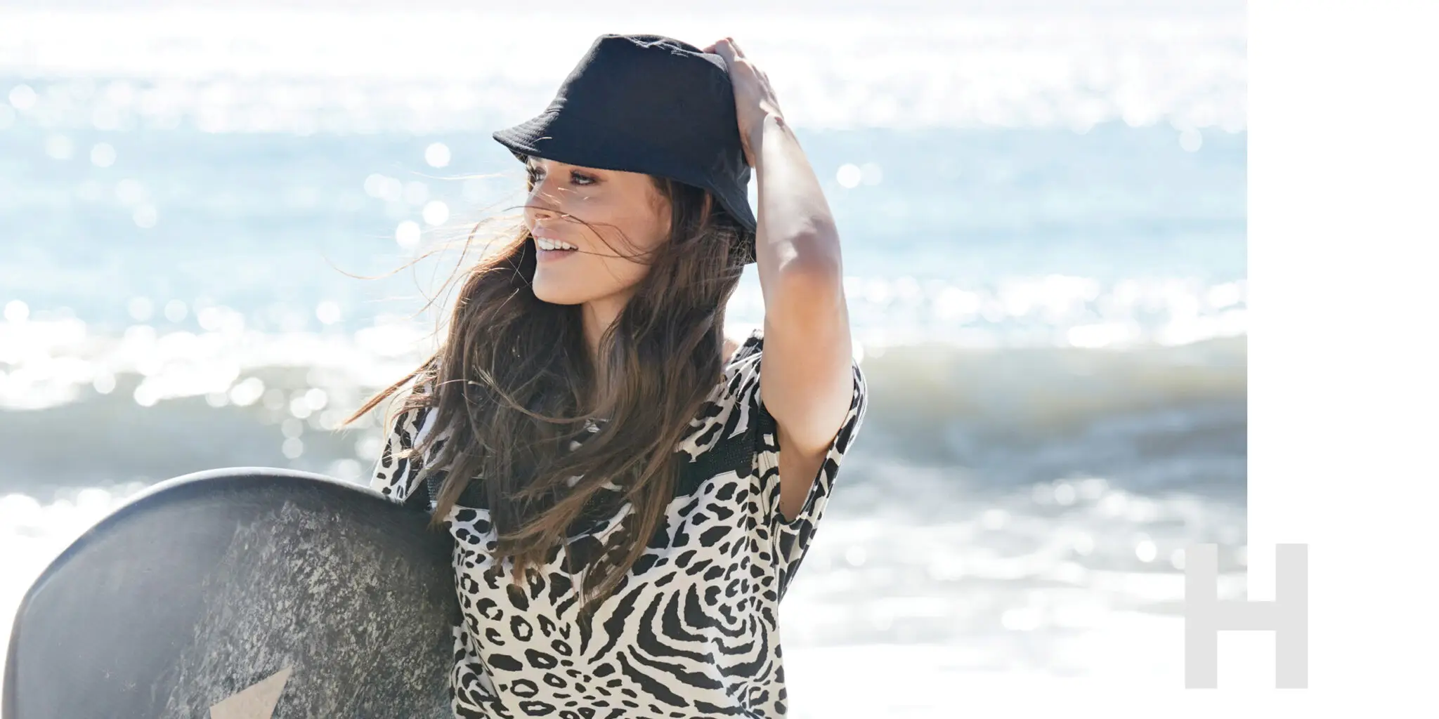 Smiling woman with long hair wearing a black bucket hat and animal print top holding a surfboard on a sunny beach.