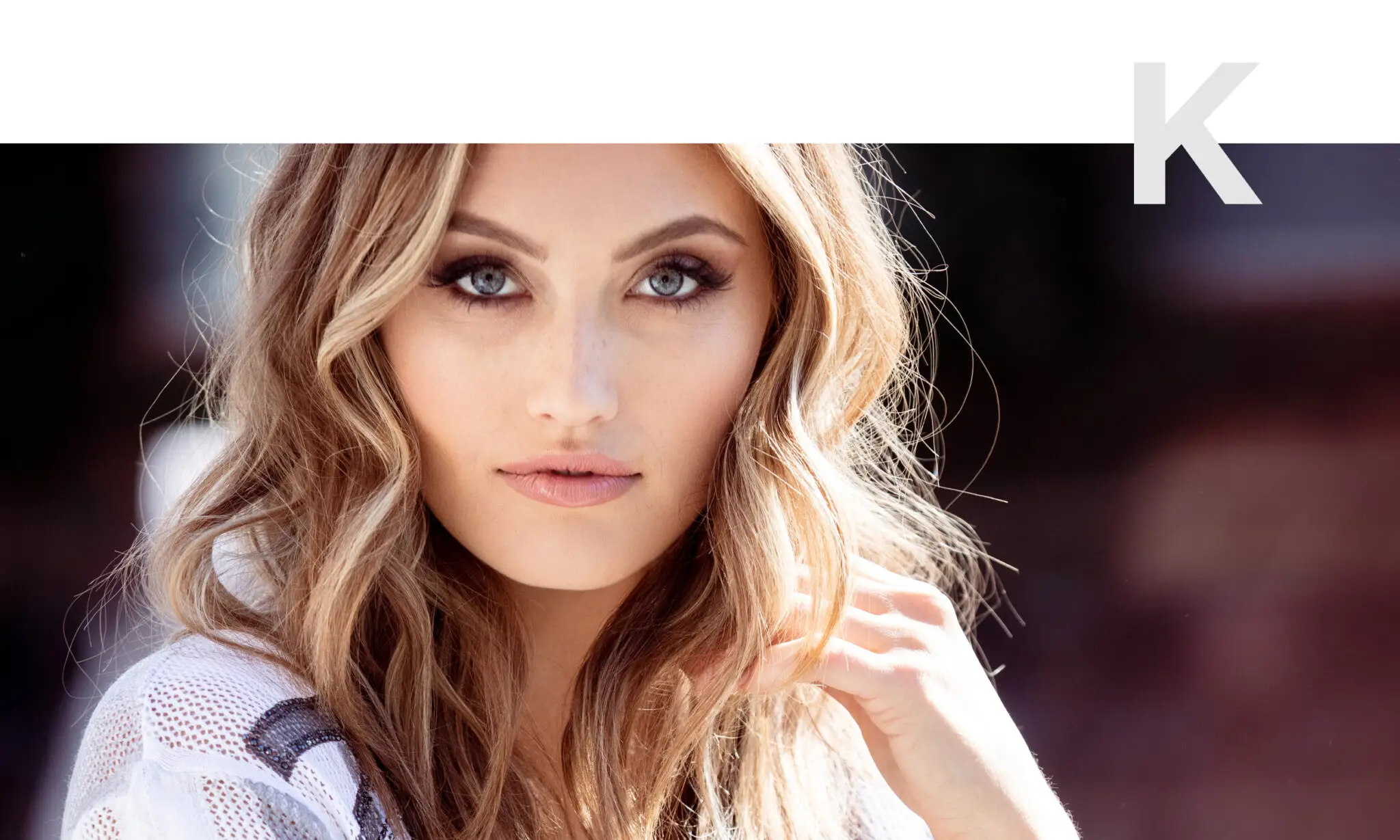 Close-up of a woman with long wavy blonde hair, blue eyes, and natural makeup looking directly at the camera.