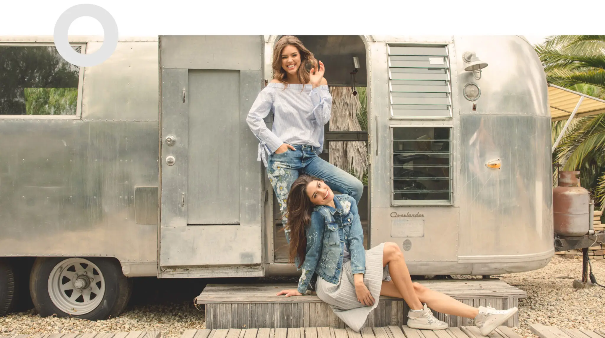 Two smiling women posing in front of a silver camper trailer, one standing in the doorway and the other sitting on wooden steps.