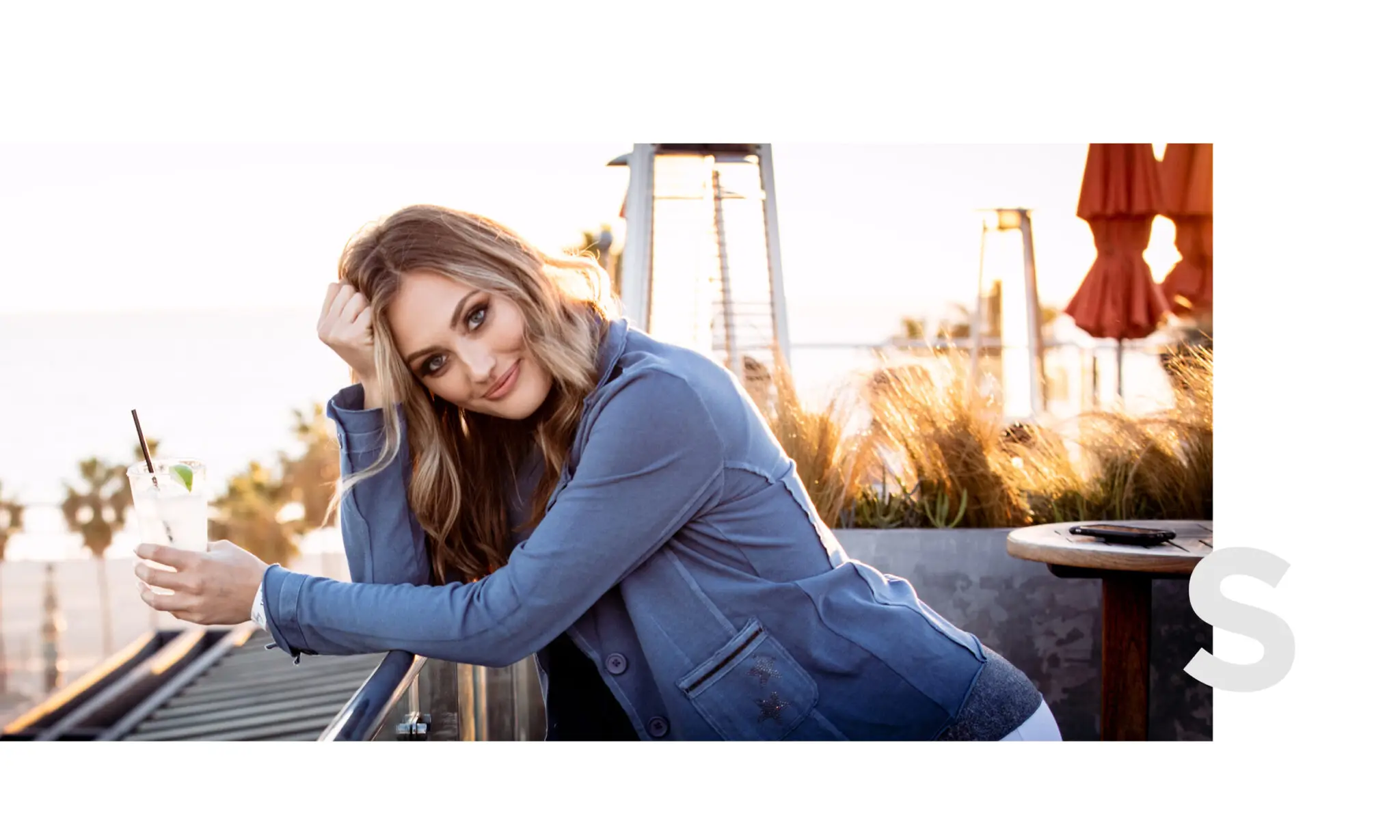 Smiling woman with light brown hair leaning on balcony railing holding a drink at sunset.