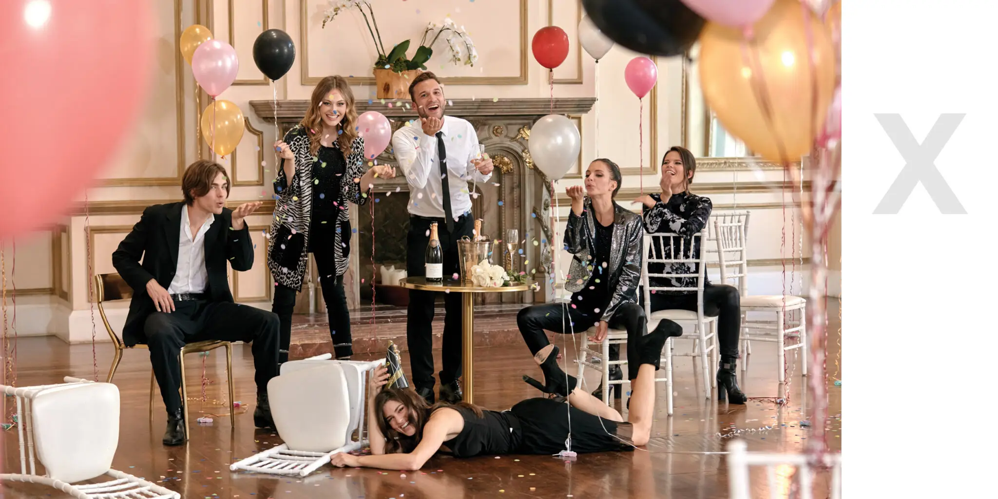 Group of six young adults celebrating indoors with balloons, confetti, champagne, and smiling faces, some sitting and one lying on the floor.