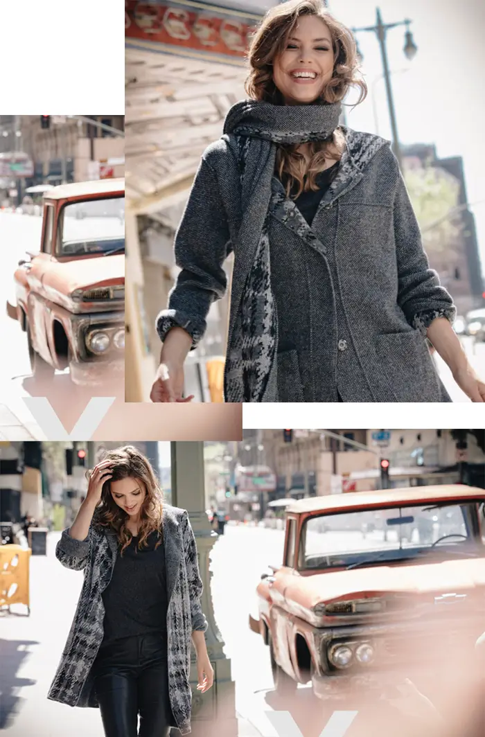 Smiling woman wearing a gray textured coat and scarf walking outdoors in city sunlight.