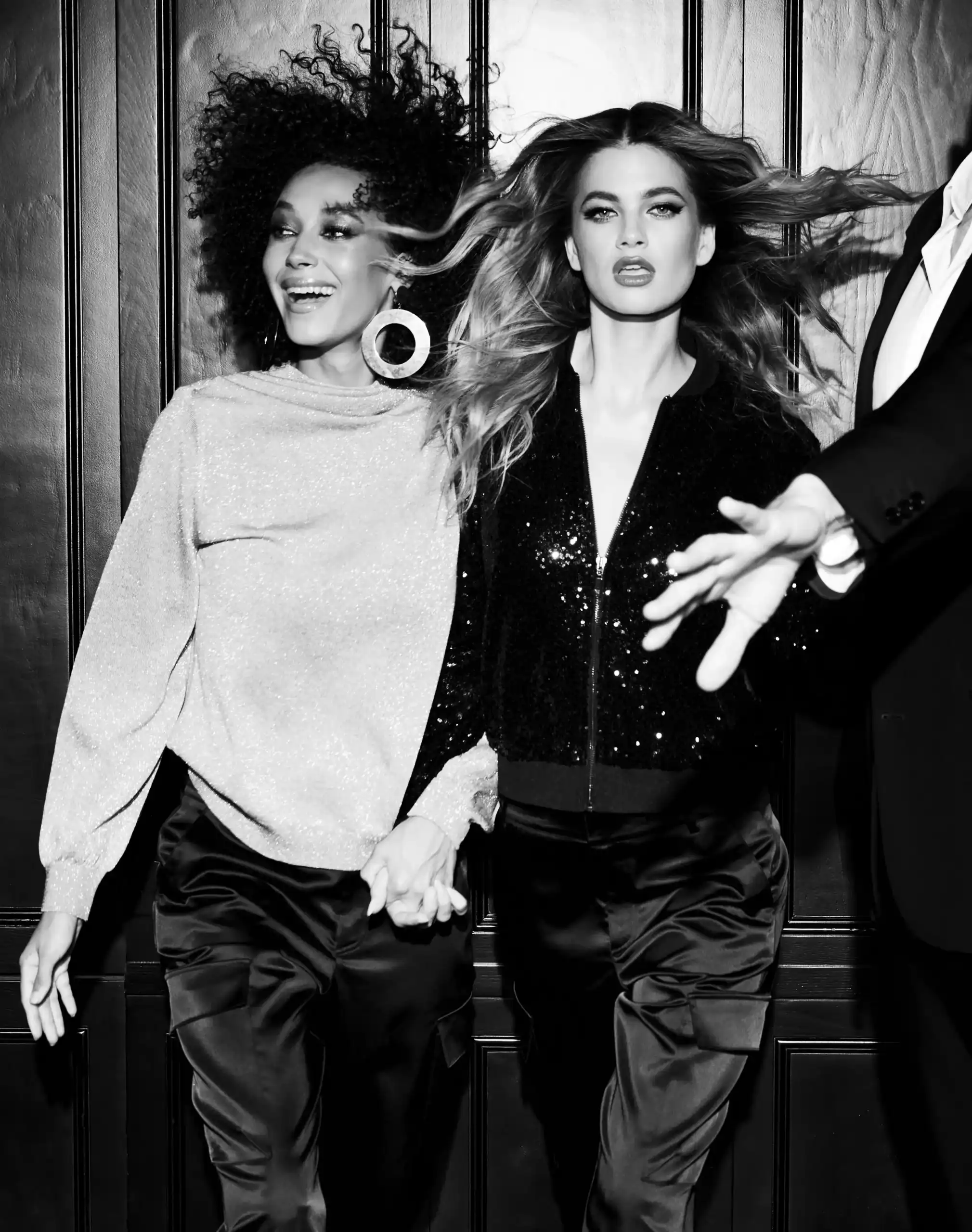 Two stylish women holding hands walking confidently with hair blowing, one smiling and the other with a serious expression.