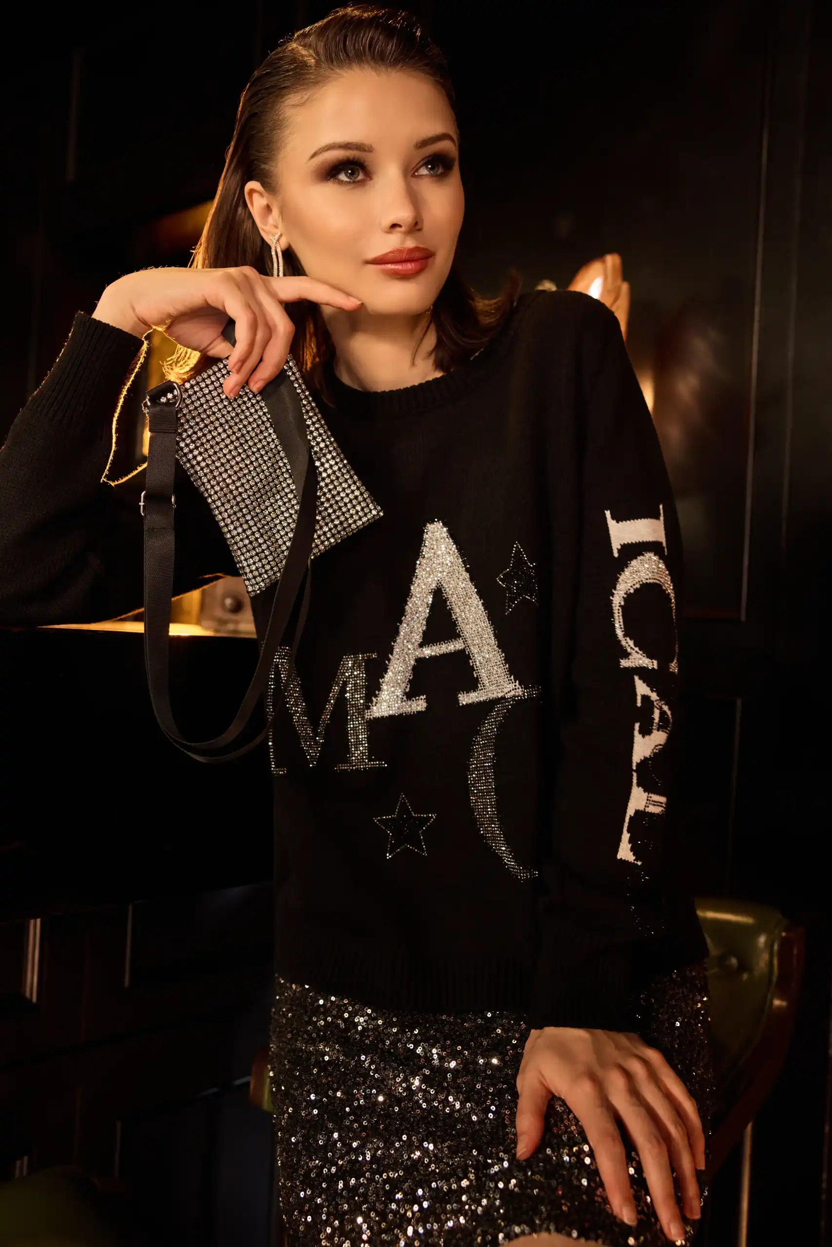 Woman wearing a black sweater with glittery silver letters and stars, holding a small rhinestone-studded purse, paired with a sparkly sequin skirt.