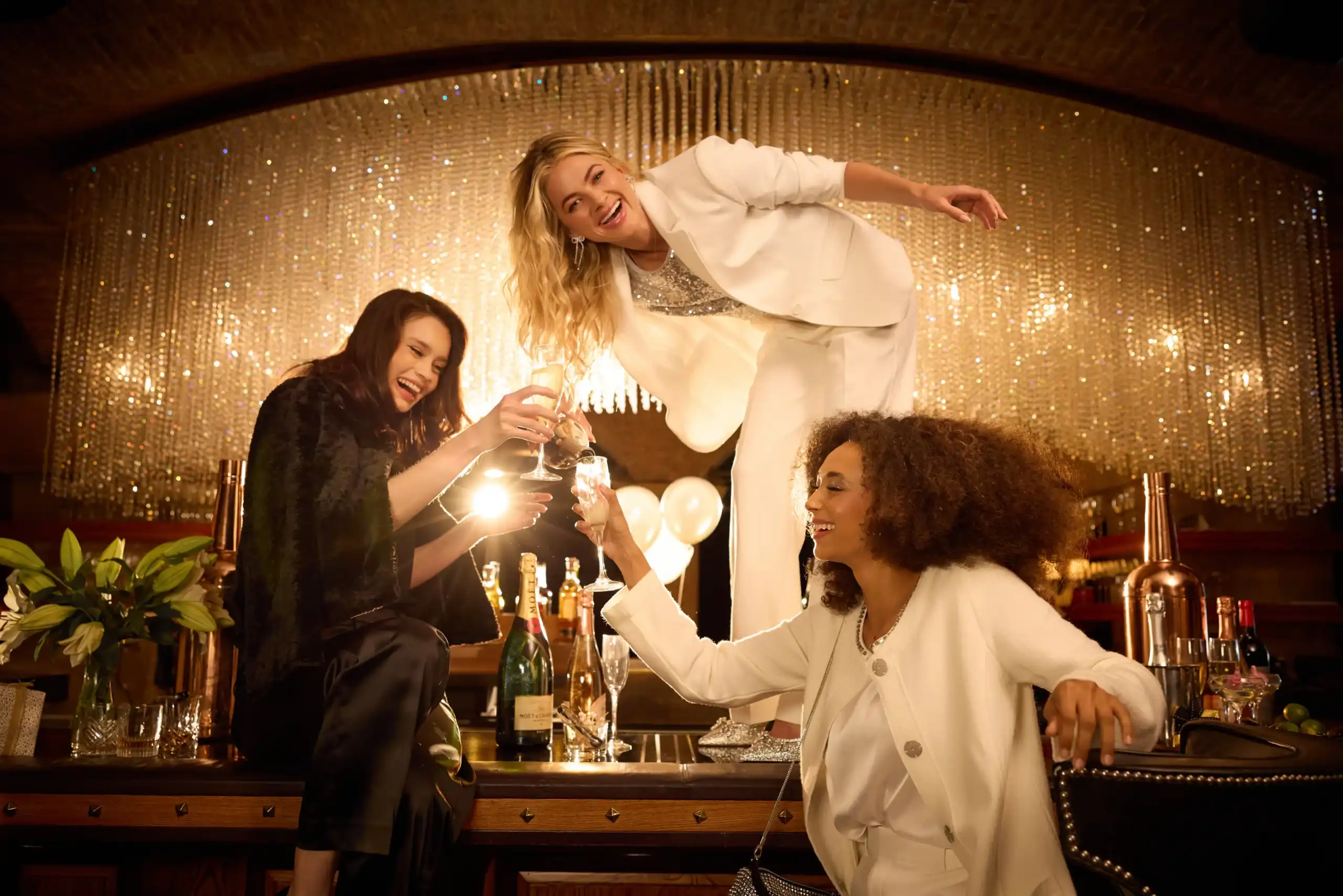 Three women dressed in elegant outfits celebrating with champagne and laughter under a large crystal chandelier.