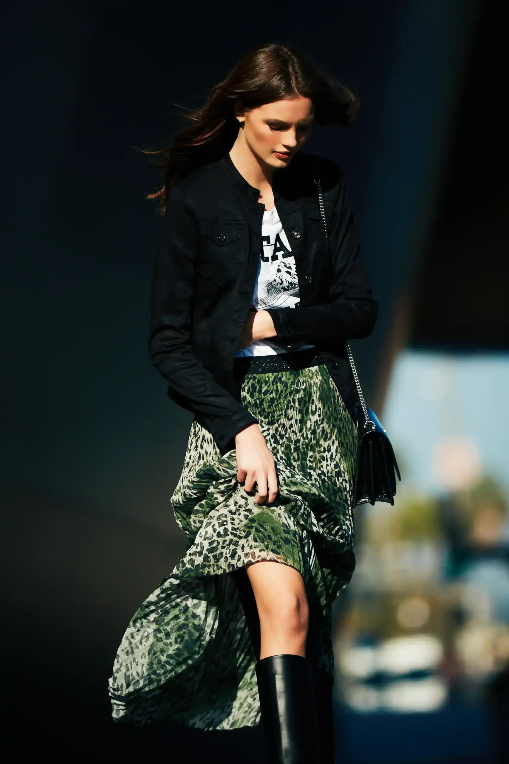 Woman wearing a black jacket, graphic white T-shirt, green leopard print skirt, and black knee-high boots walking outdoors.