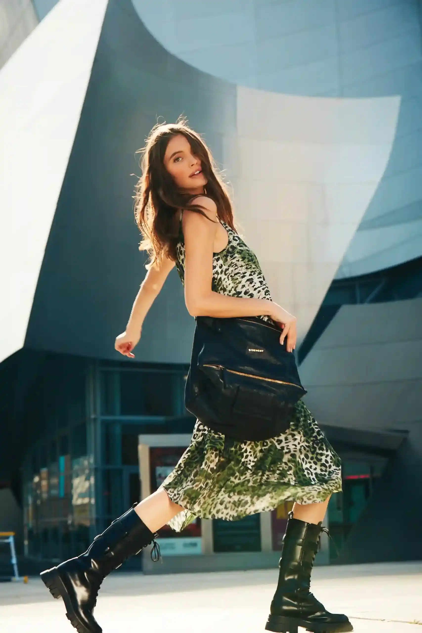 Woman walking outside wearing a green leopard print dress, black knee-high boots, and carrying a black Givenchy bag.