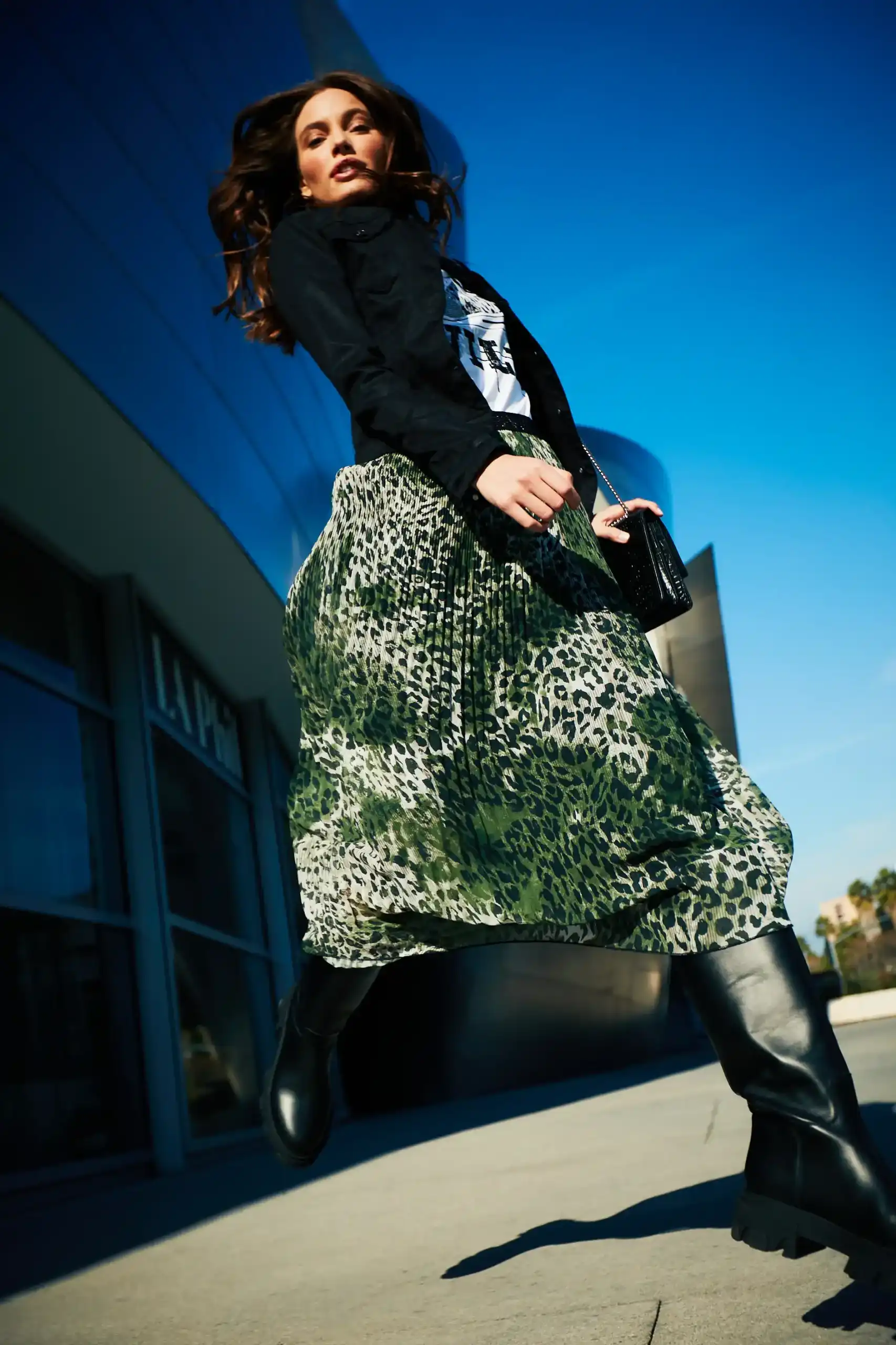 Low-angle view of a woman wearing a green leopard print skirt, black jacket, and black boots, walking outdoors against a blue sky.