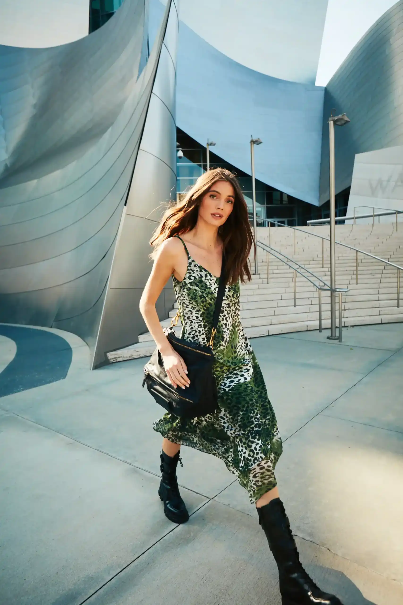 Woman walking outdoors in a green and white animal print dress with black boots and a black bag, against a backdrop of modern metallic architecture and stairs.