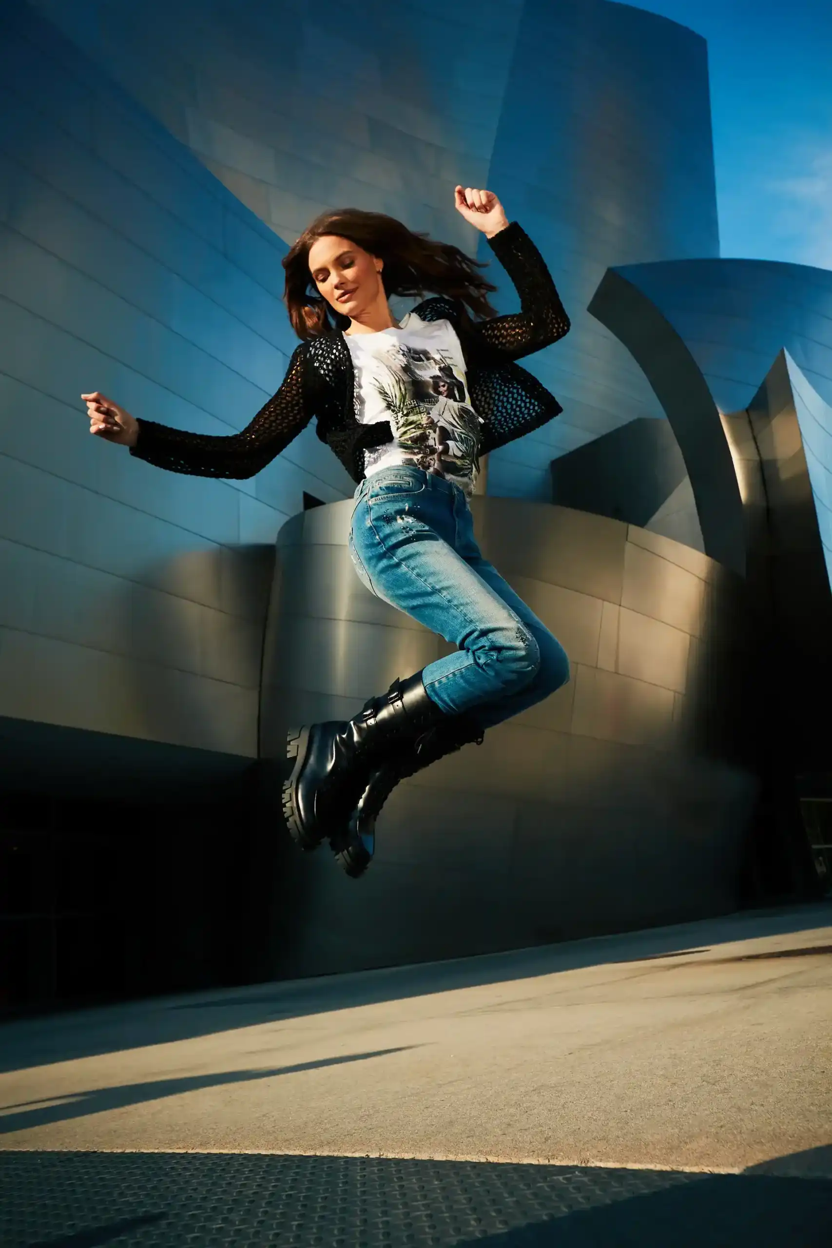 Woman in casual clothes and black boots jumping with a modern metallic building in the background.