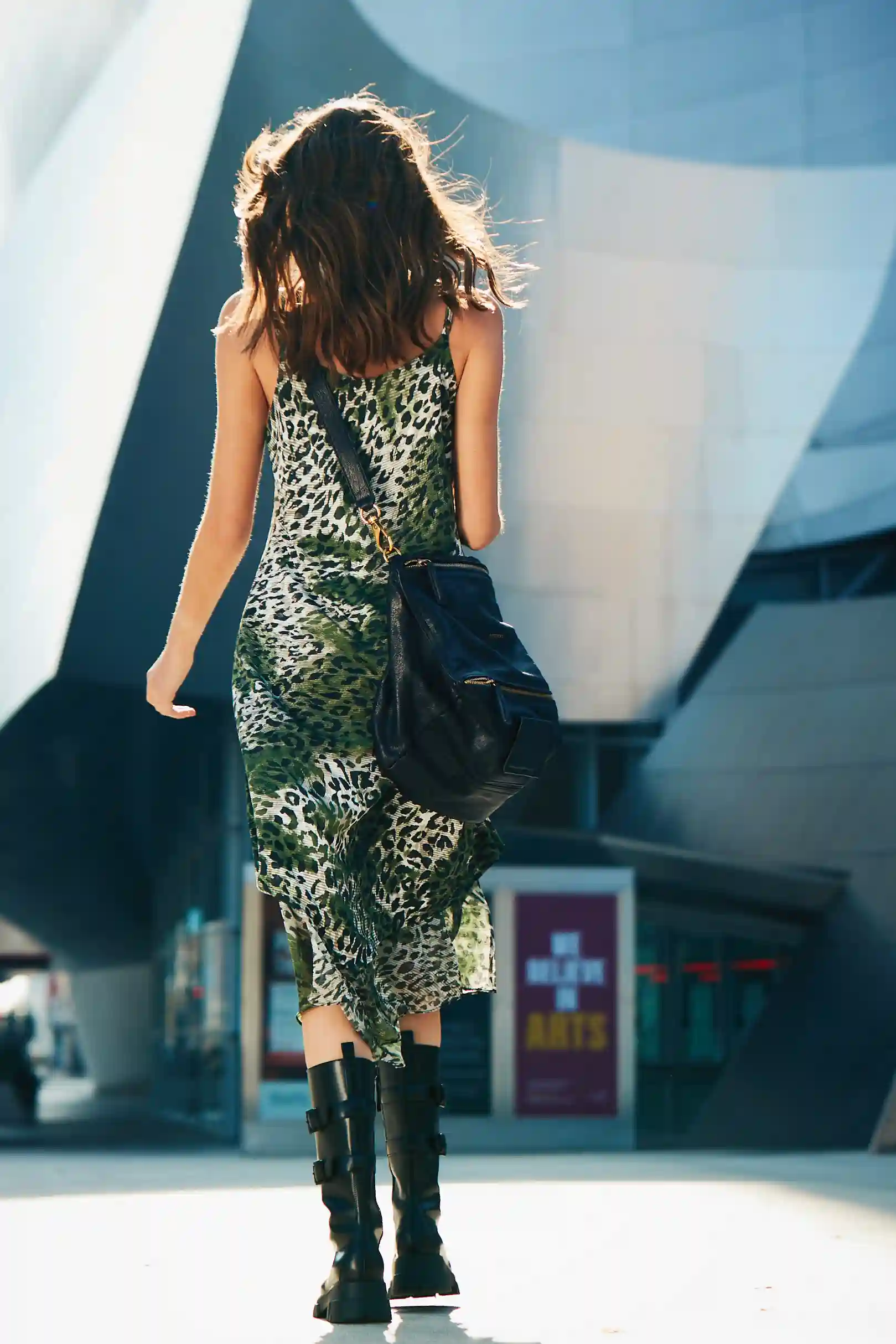 Woman walking away in a green leopard print dress with black boots and a black shoulder bag in an urban setting.