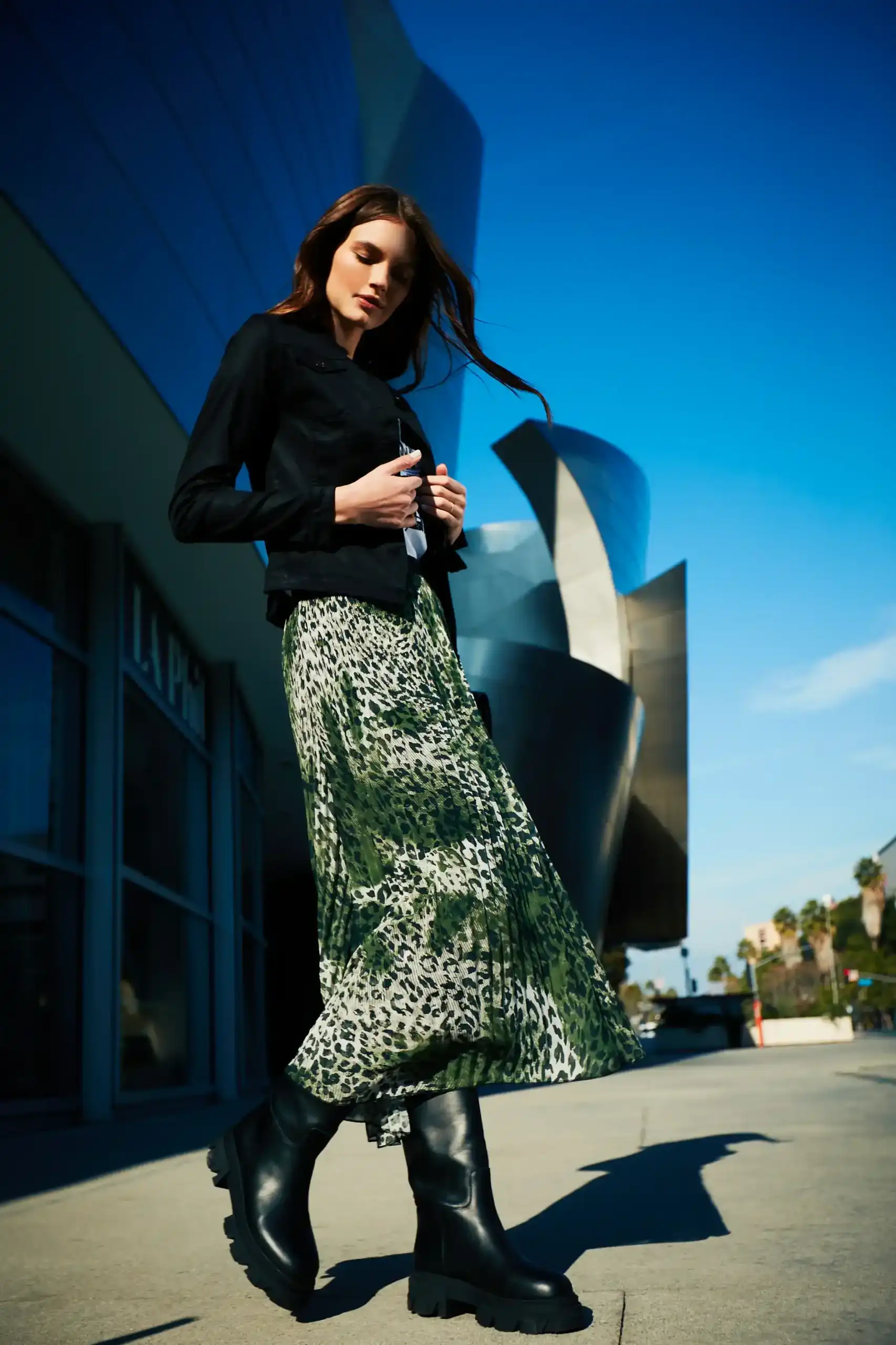 Woman wearing a black jacket, green leopard print skirt, and black boots walking outdoors with modern architecture and clear blue sky in the background.