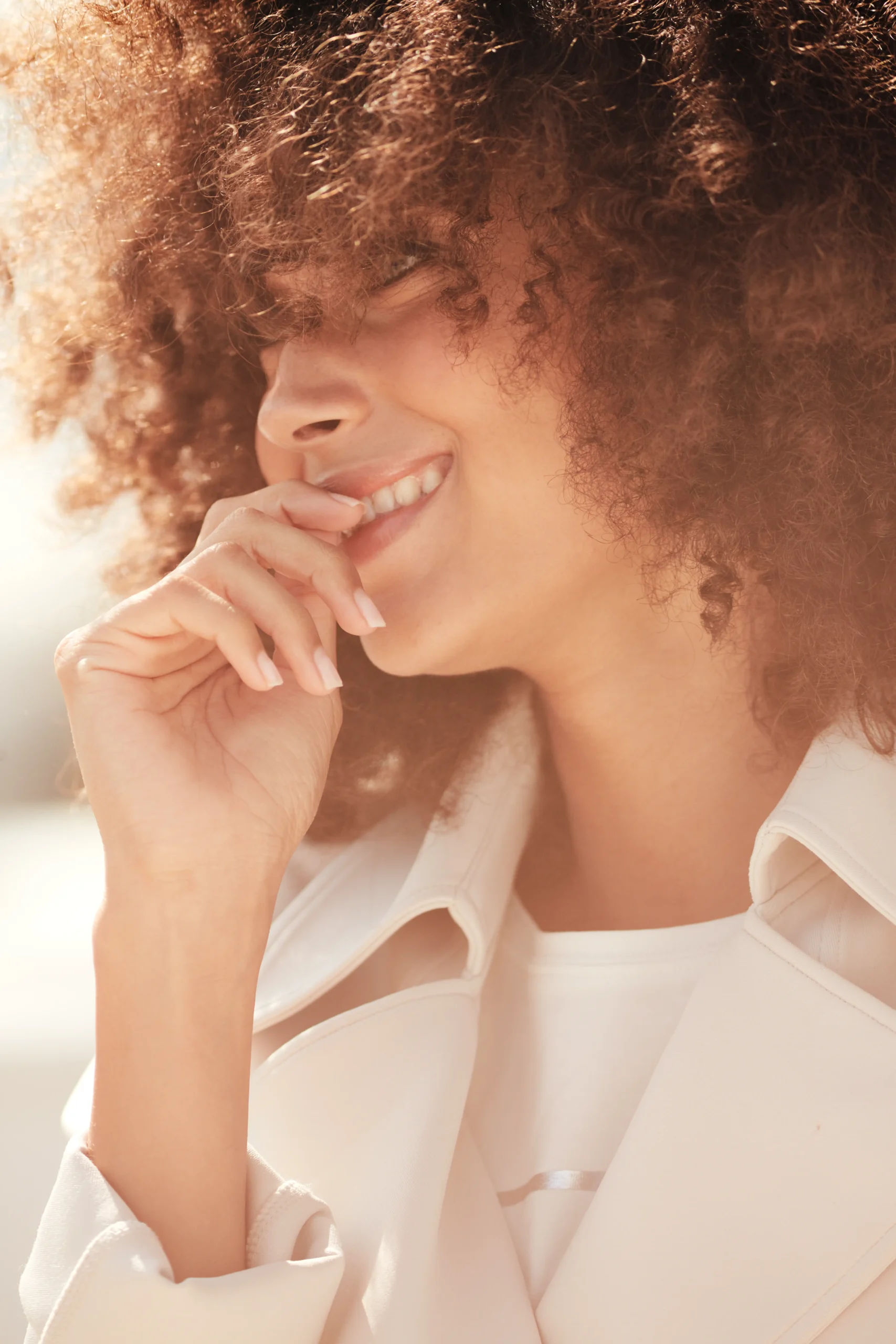 Close-up of a smiling woman with curly hair covering part of her face, wearing a light-colored jacket, with her hand near her mouth.