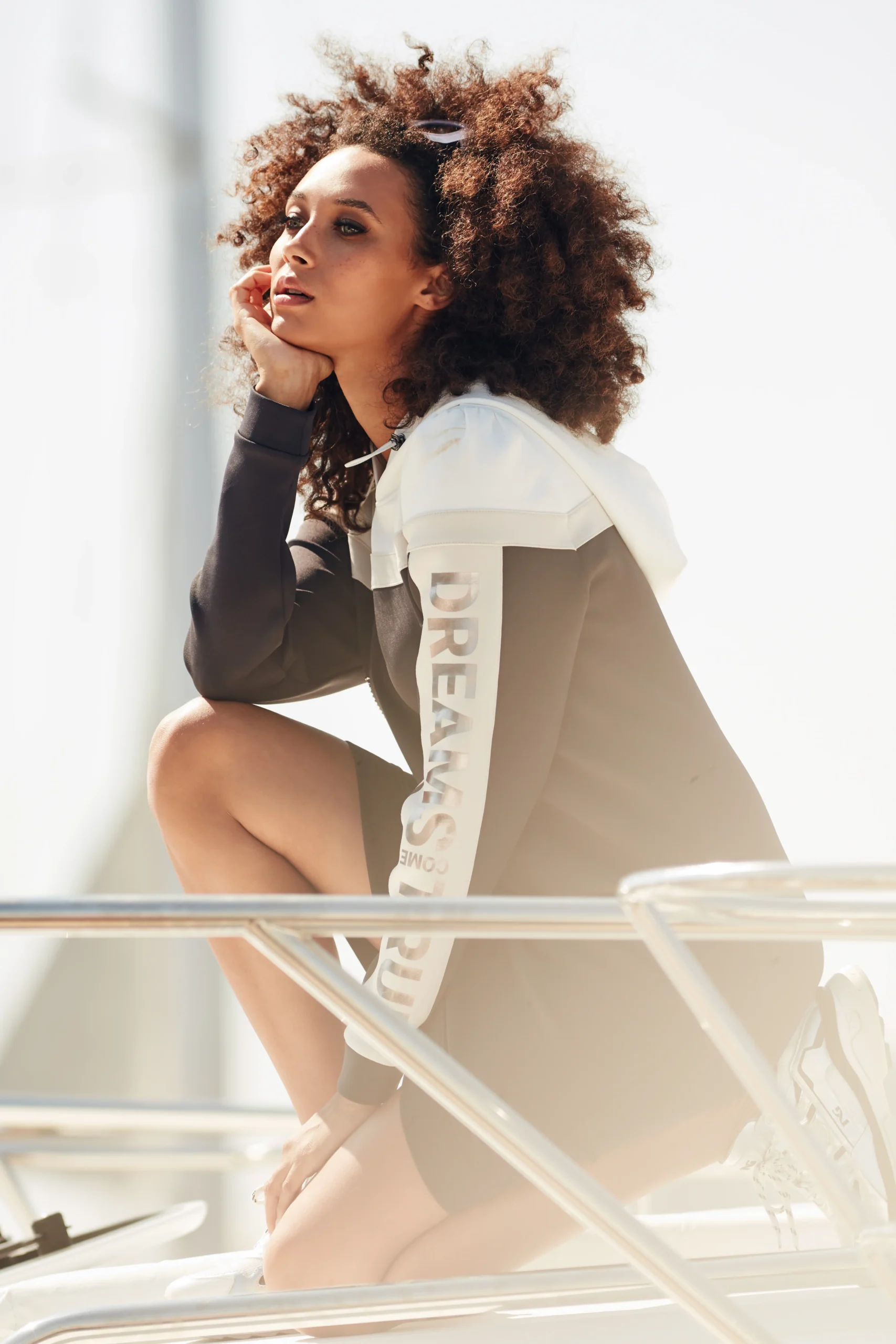 Woman with curly hair in a black and white hoodie crouching on a boat railing in bright sunlight.