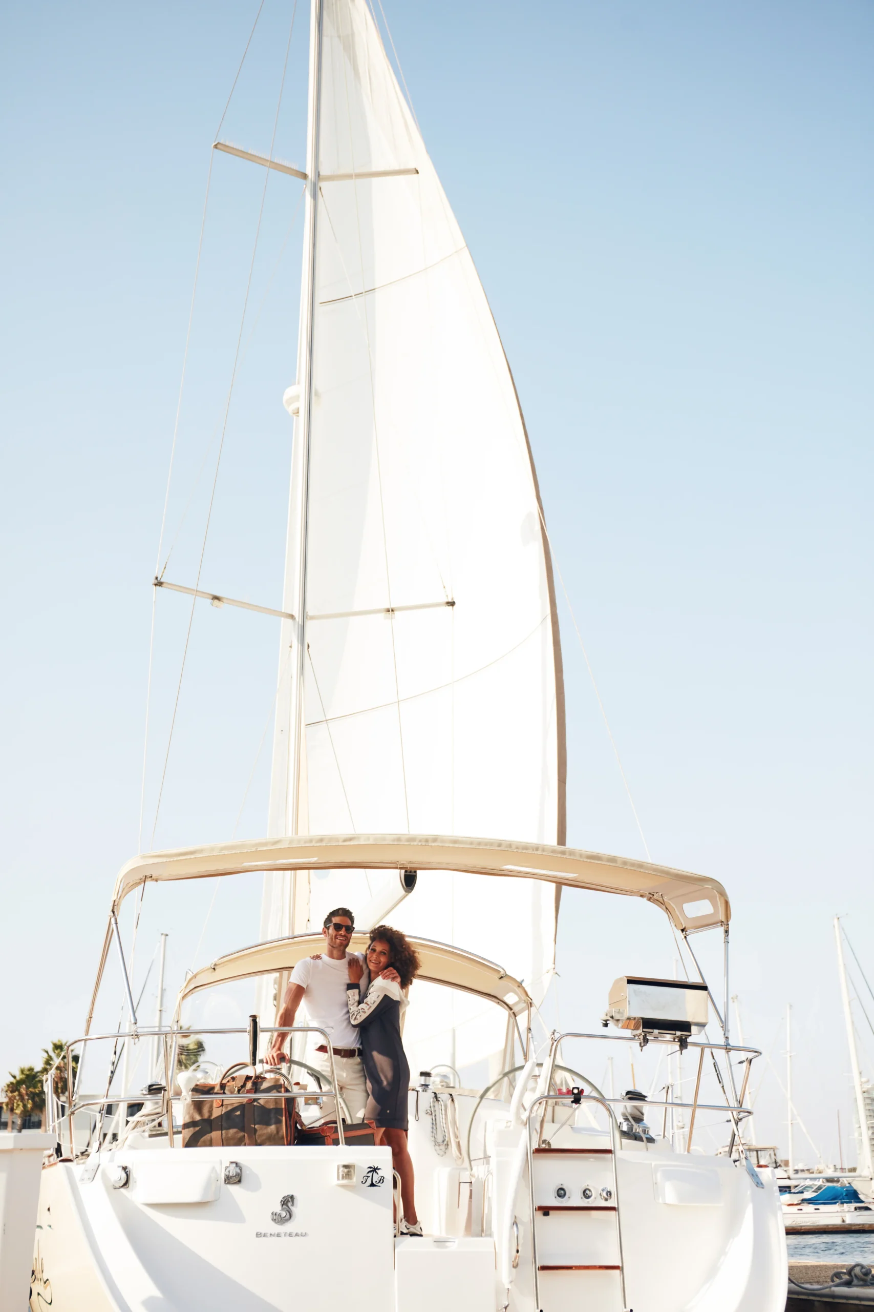 Smiling couple standing on the deck of a white sailboat docked at a marina under clear sky.