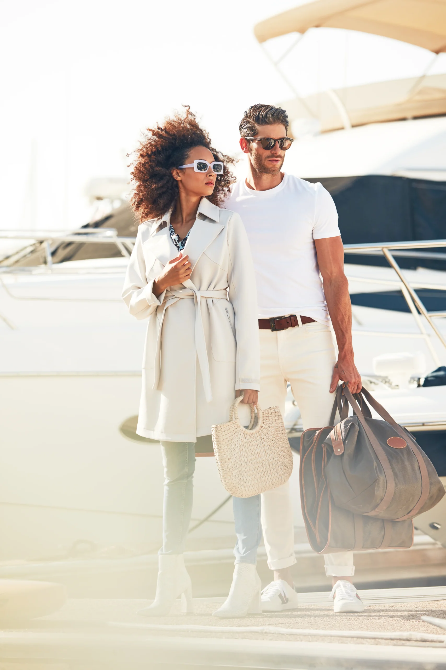 Fashionable couple wearing sunglasses standing on a dock by a yacht, holding a woven handbag and travel bags.