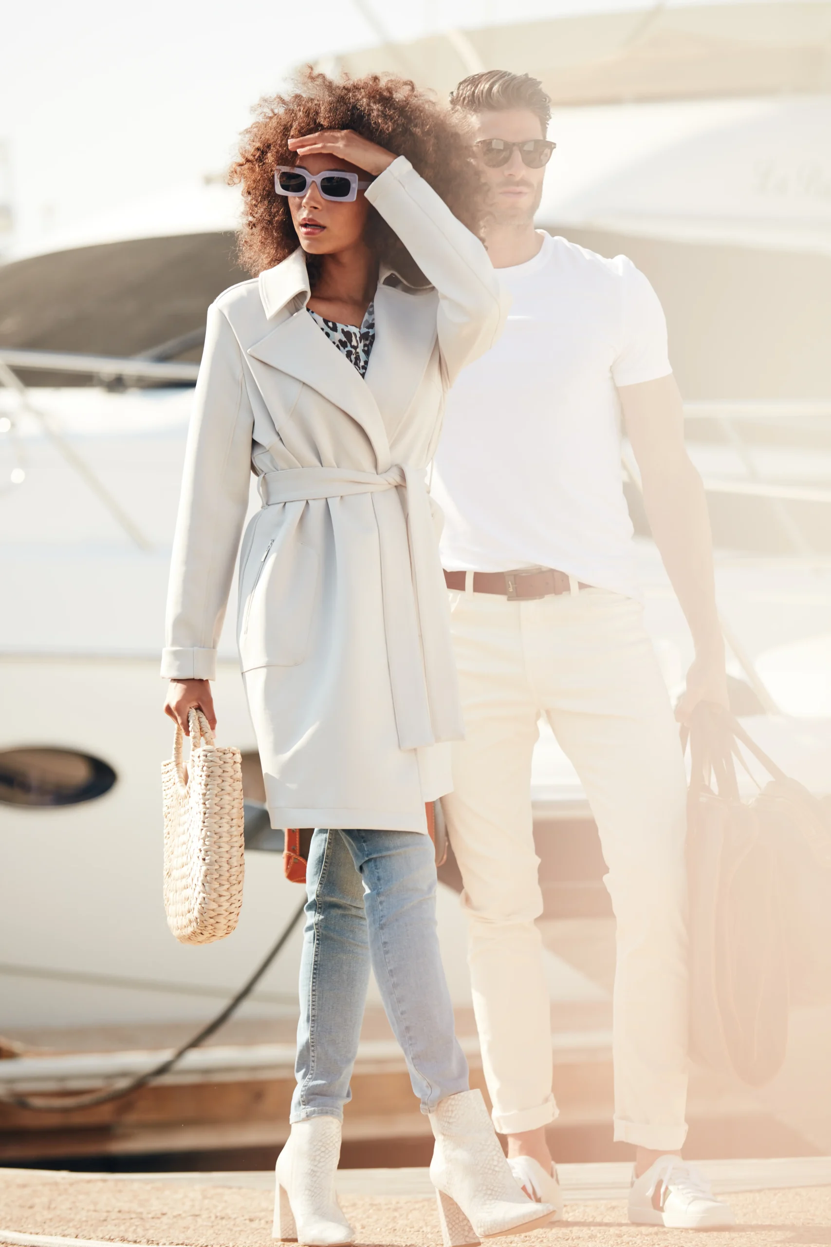 Fashionable woman in sunglasses and a white coat holding a woven bag standing next to a man in white shirt and pants near a yacht.