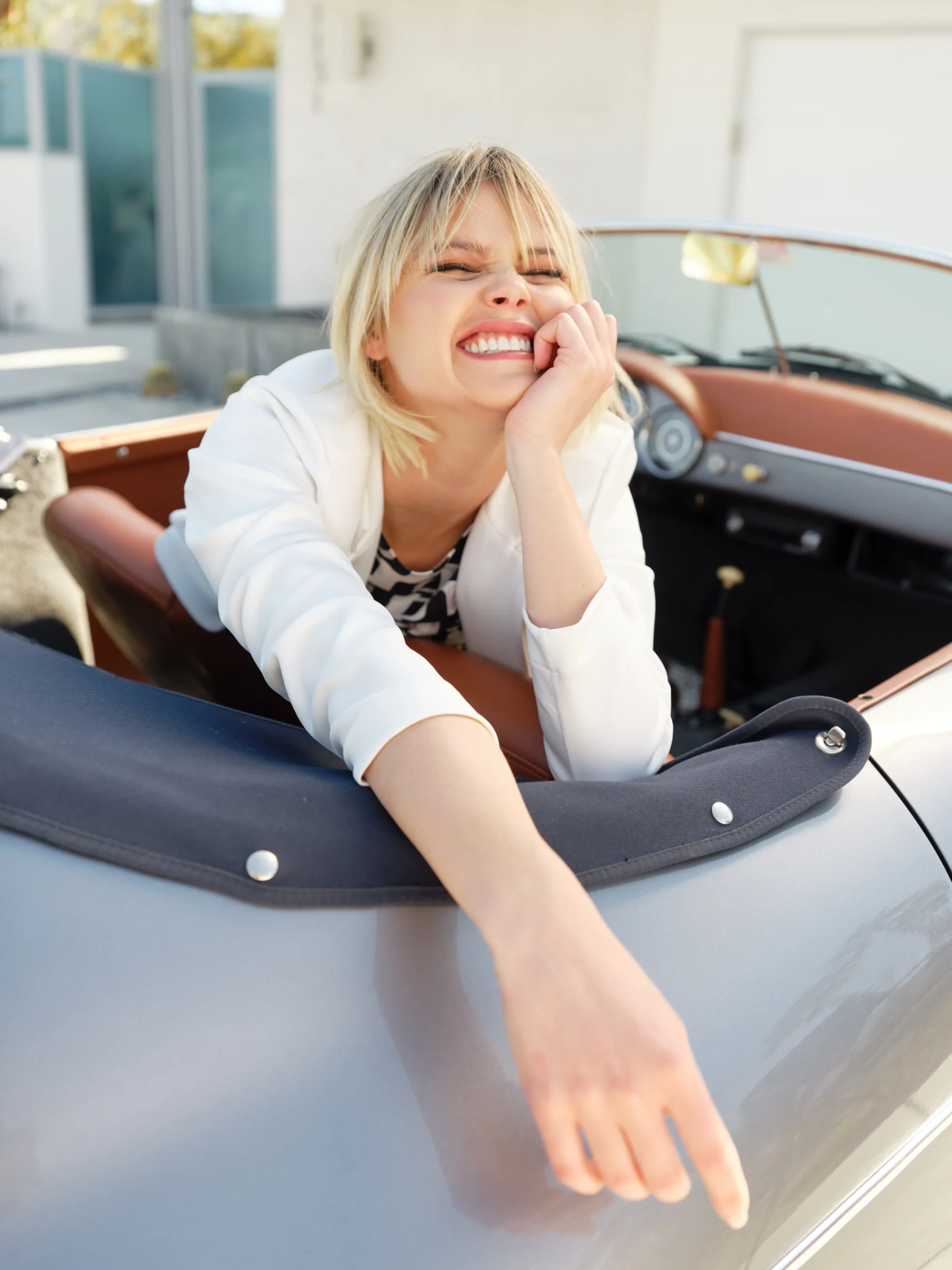 Smiling blonde woman in white jacket leaning out of a silver convertible car.