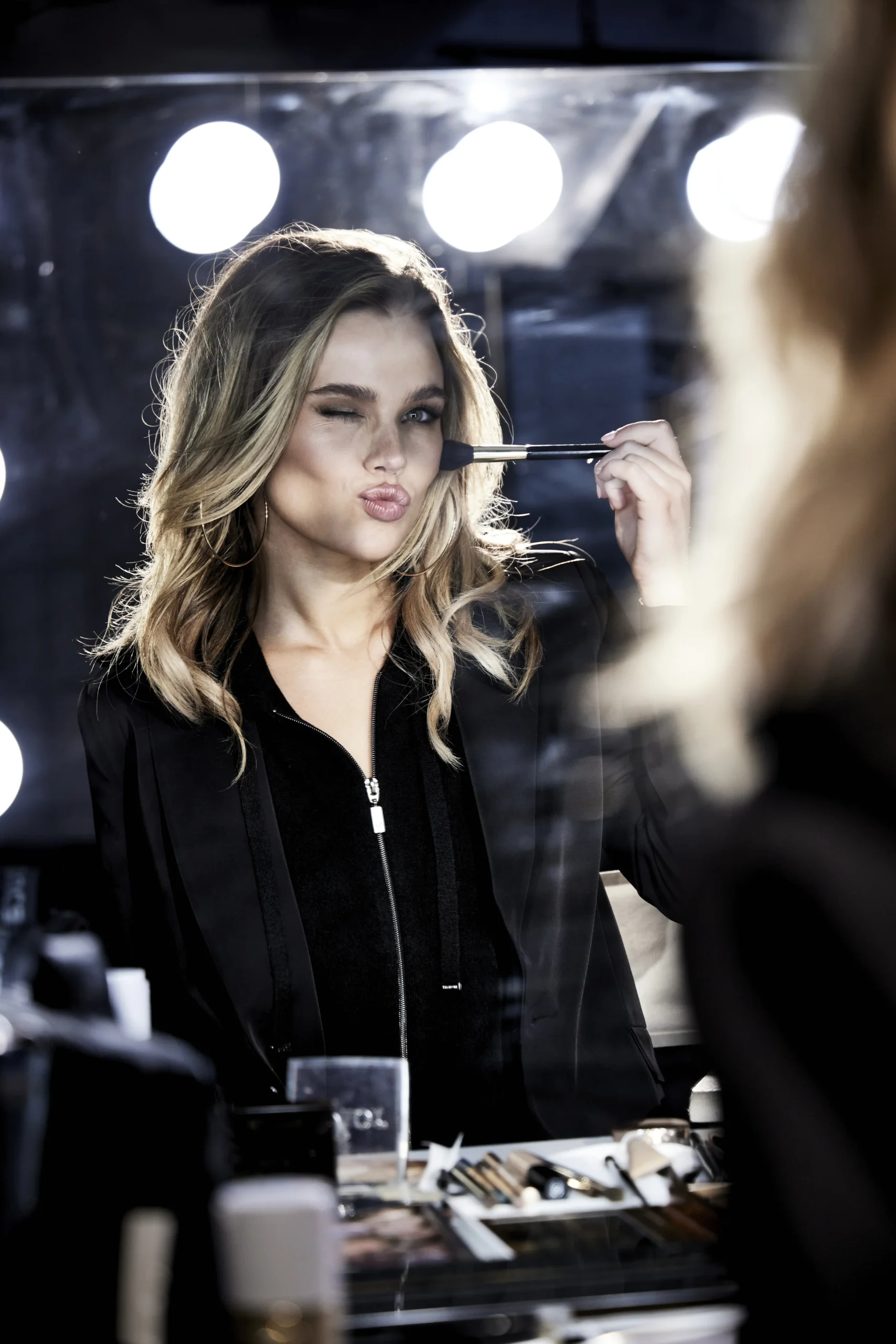 Blonde woman with wavy hair applies makeup with a brush while winking in front of a vanity mirror surrounded by lights.