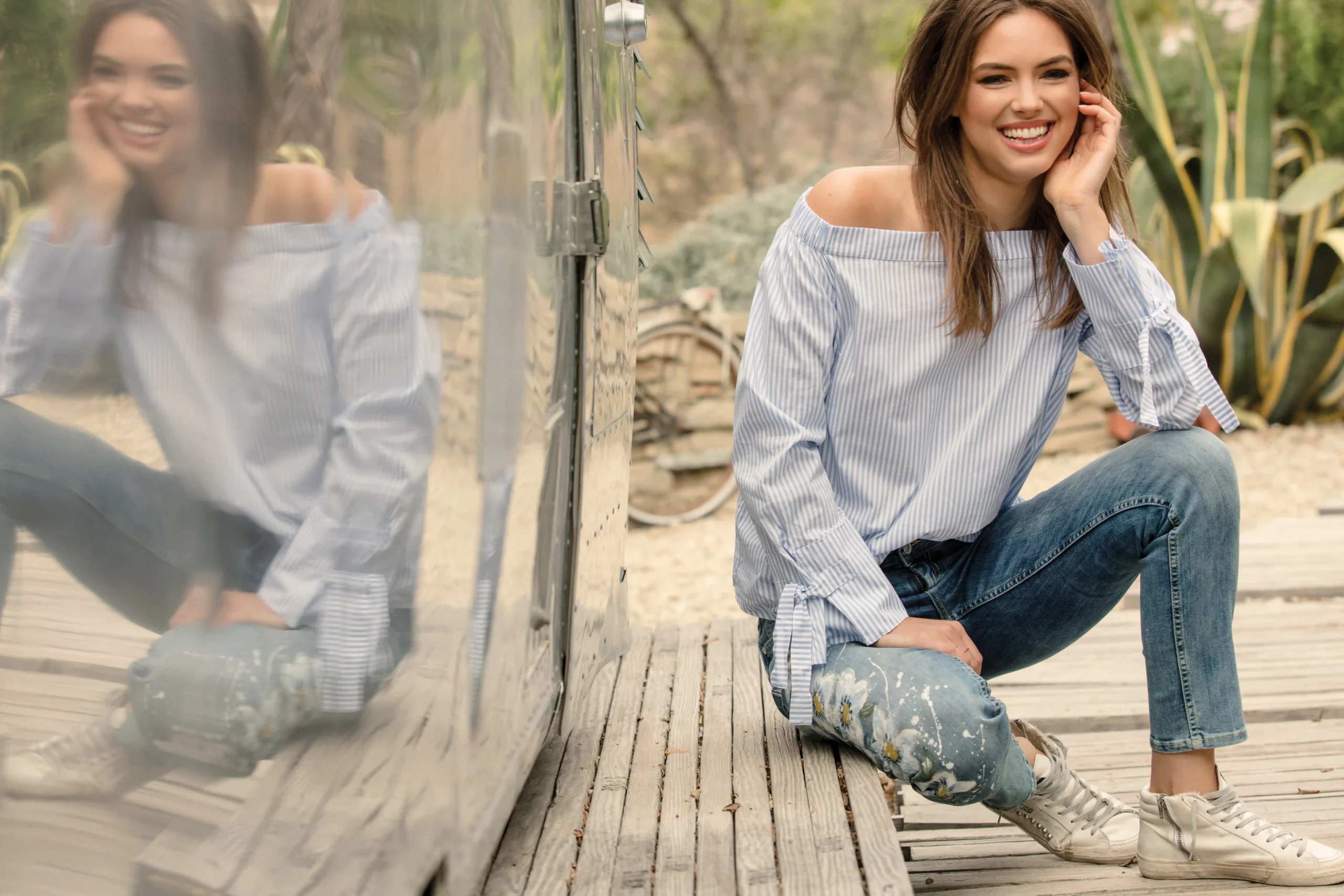 Smiling woman wearing an off-the-shoulder striped blouse and painted jeans sitting on wooden decking next to a reflective metallic surface.