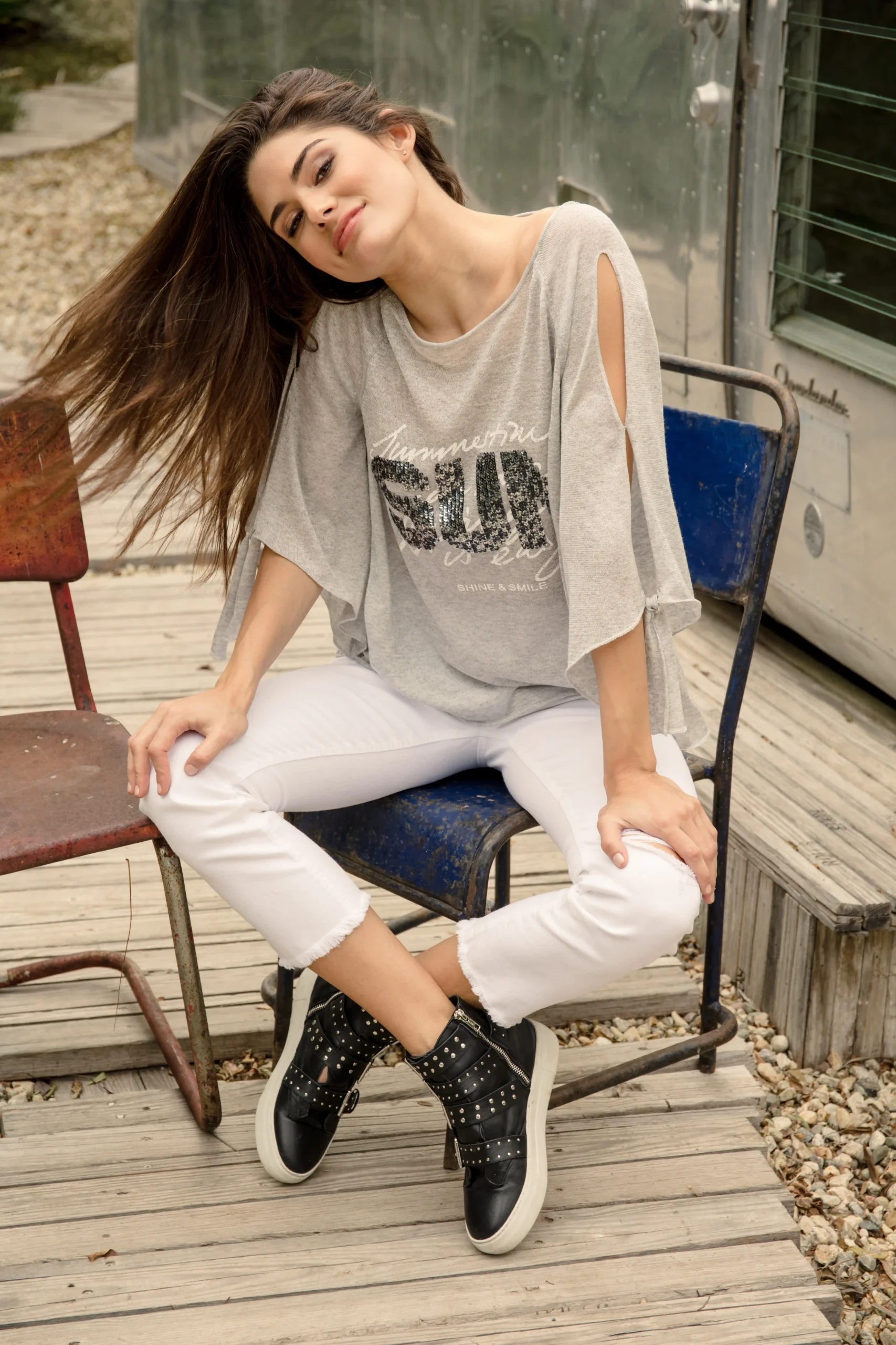 Woman with long brown hair sitting on a rustic blue metal chair, wearing a gray top with cut-out sleeves, white cropped pants, and black studded sneakers.