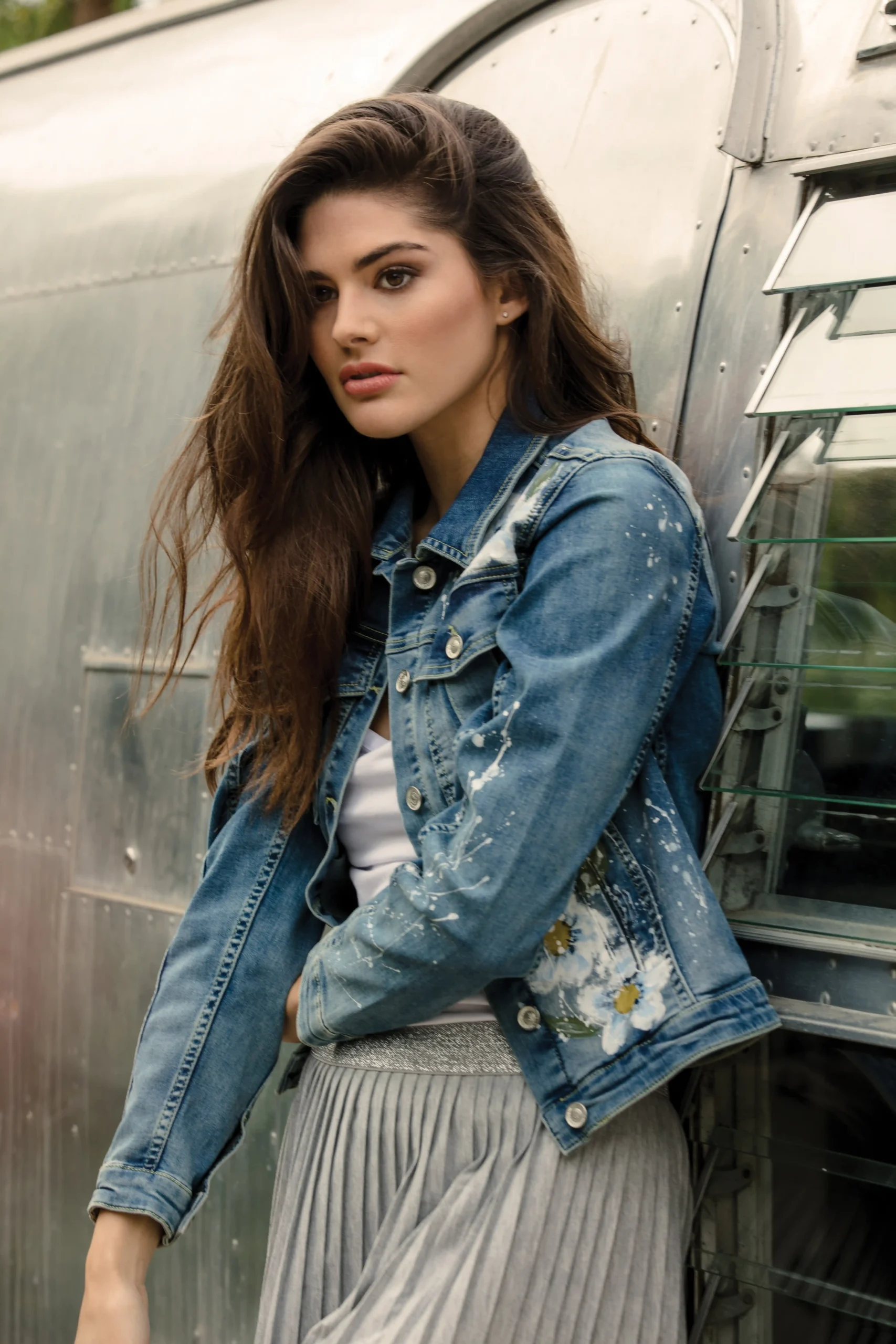 Young woman with long brown hair wearing a denim jacket with floral design leaning against a metallic surface.