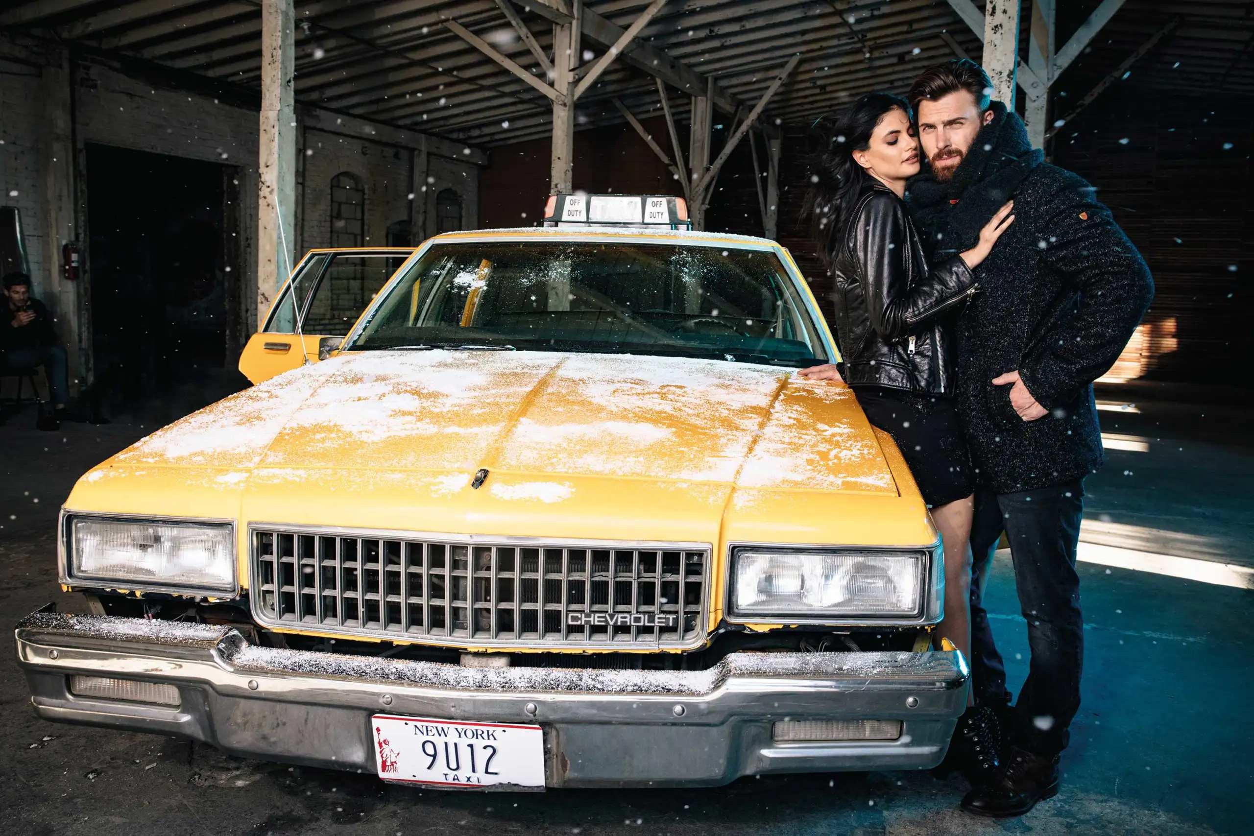 Couple in winter clothing embracing beside a snow-dusted yellow New York taxi inside a dim warehouse.