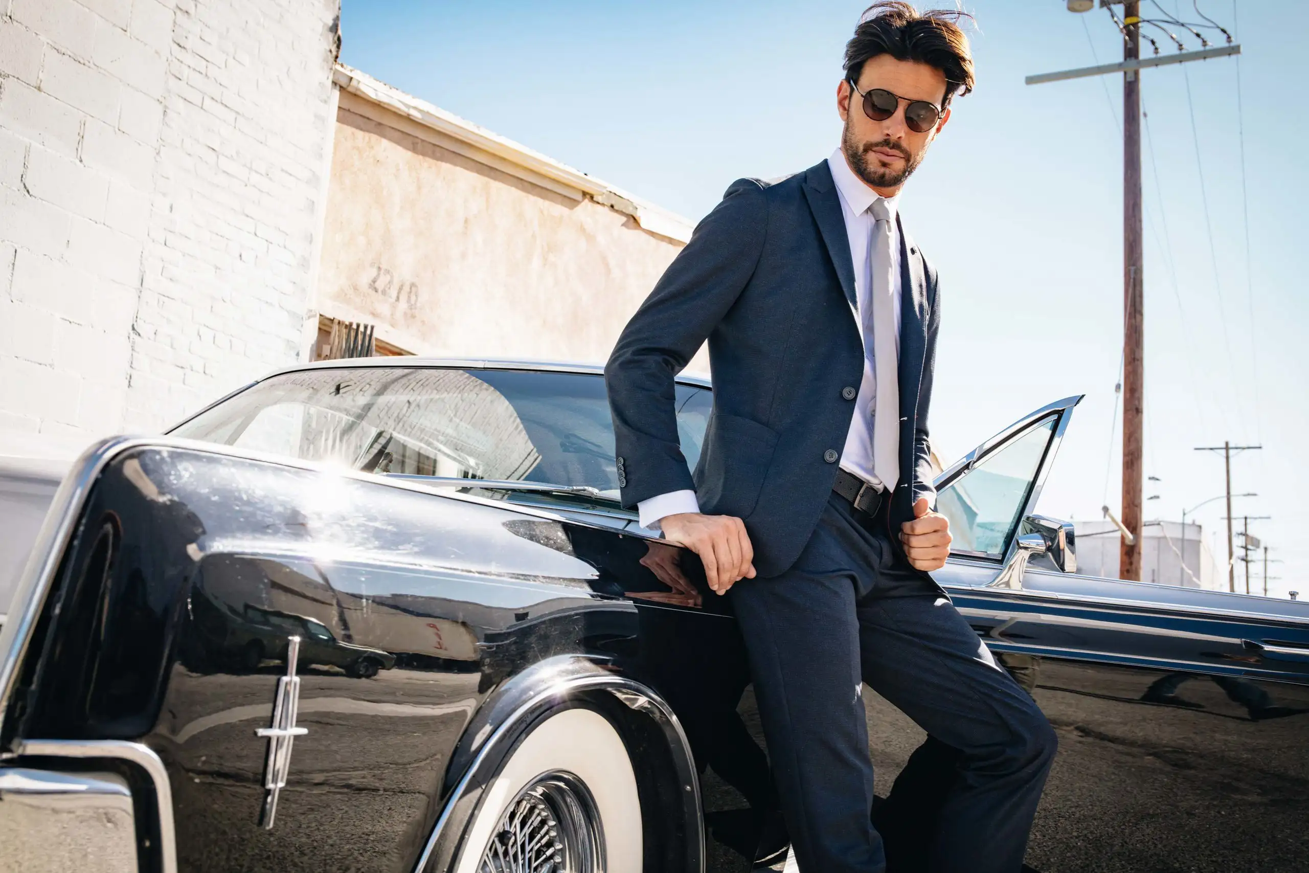 Man in a dark suit and sunglasses leaning against a classic black car with whitewall tires on a sunny day.