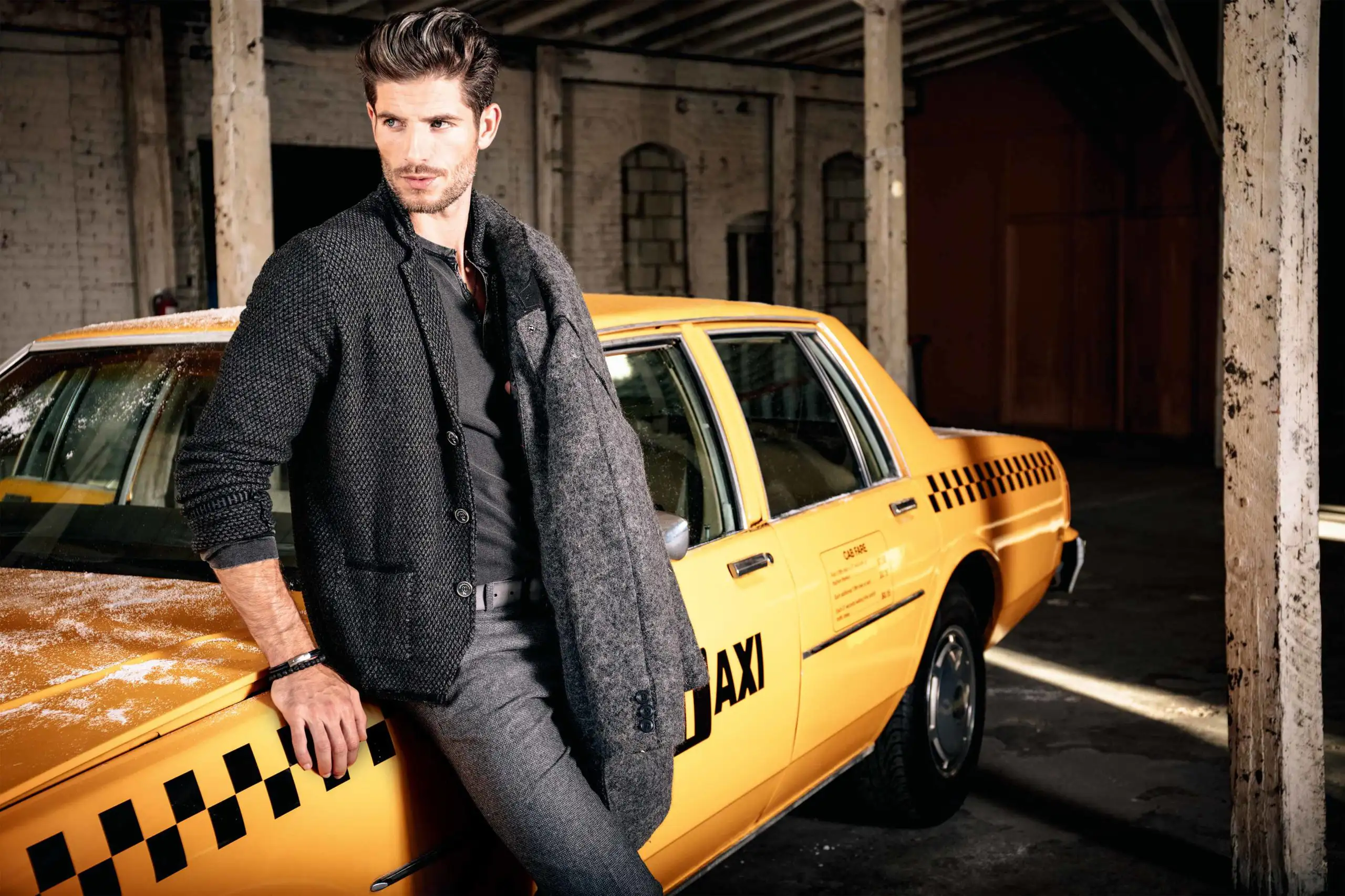 Man in dark textured jacket leaning on a yellow taxi inside a dimly lit garage.