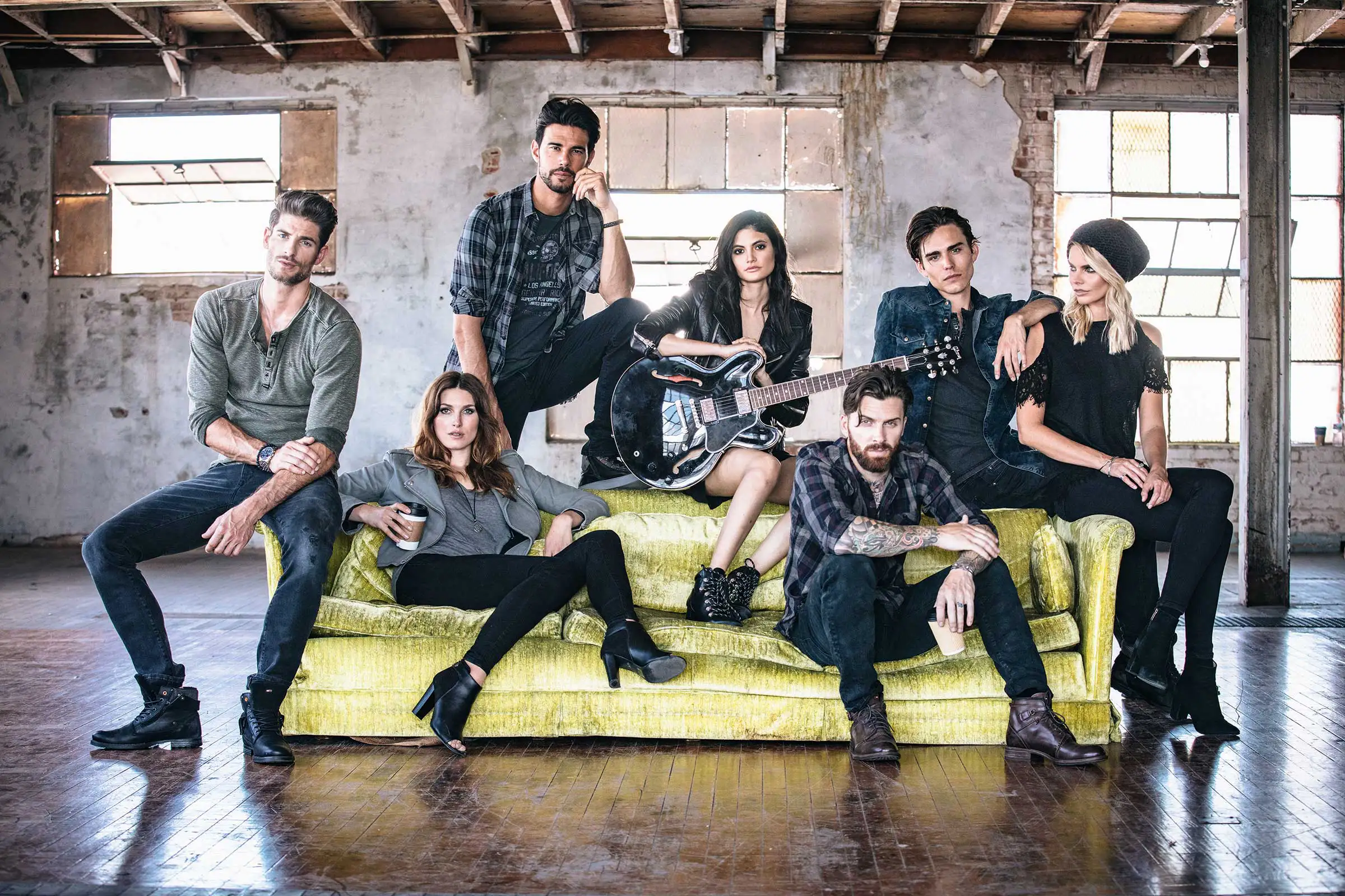 Group of seven young adults posing on and around a yellow couch in a rustic room, with one woman holding a black electric guitar.