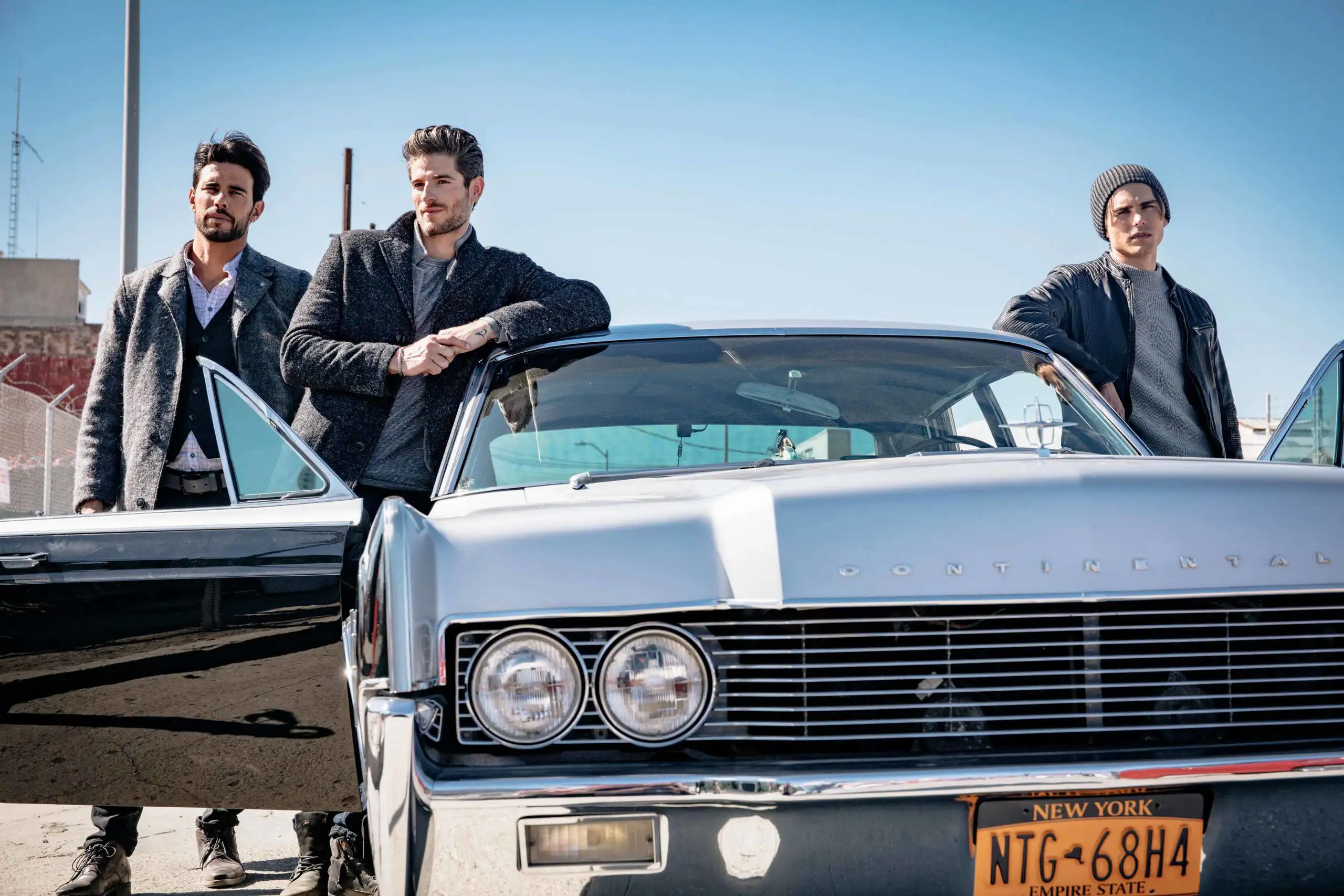 Three men posing with a classic silver Lincoln Continental car with a New York license plate under a clear blue sky.