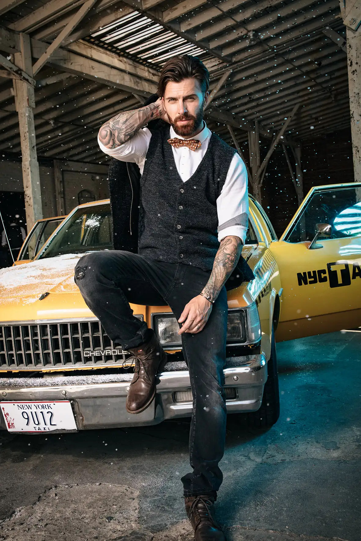 Tattooed man with beard wearing a bow tie and vest, sitting on the front bumper of a yellow NYC taxi with the driver door open inside a garage.