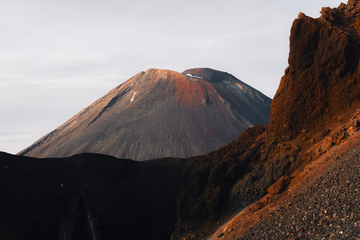 Sunlit volcanic mountain with rocky slopes and a crater under a cloudy sky.