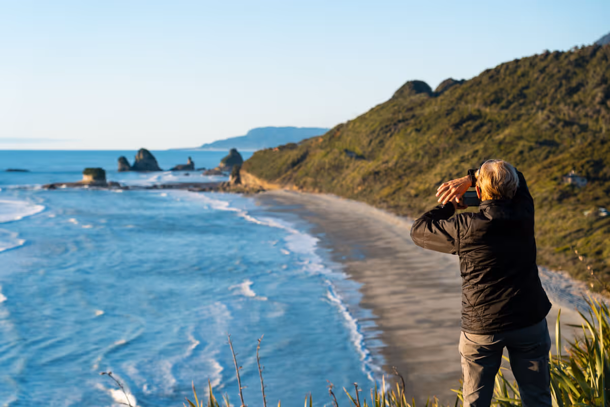 Person in a black jacket taking a photo of a scenic beach with rocky hills and waves.