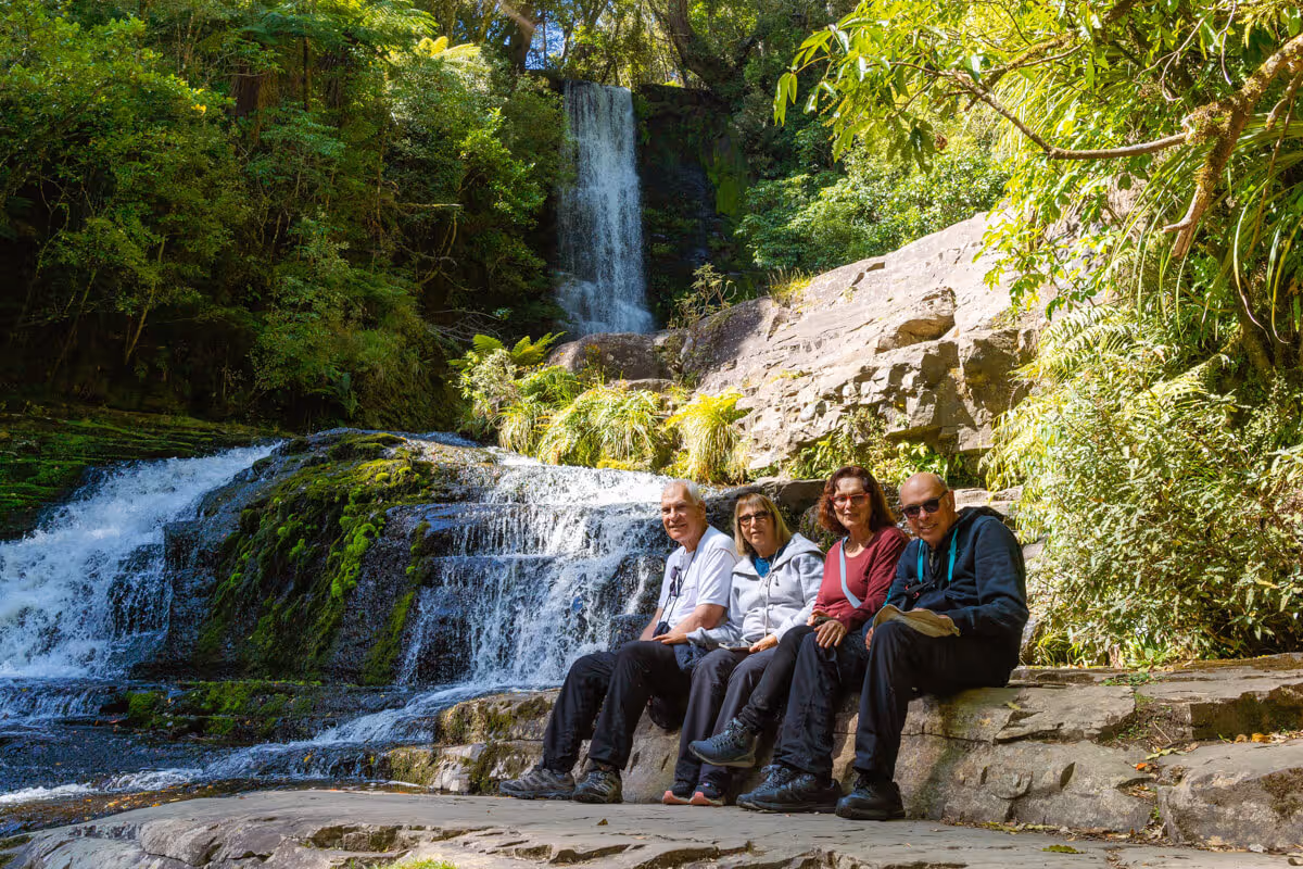 Four people sitting on rocks near a flowing waterfall surrounded by lush green vegetation in a forest.