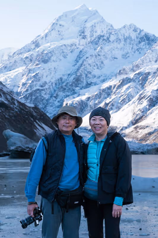 A smiling couple dressed in winter clothing stands together in front of a snowy mountain landscape, with a camera in the man's hand.