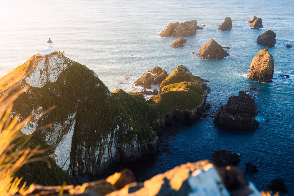 Lighthouse atop a rugged cliff surrounded by rocky islets in blue ocean water at sunset.
