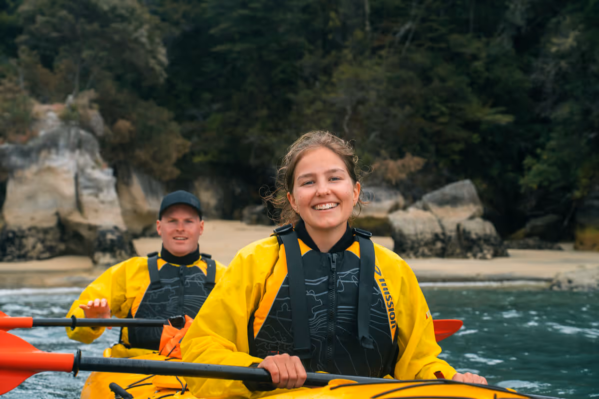 Smiling woman and man wearing yellow jackets kayaking on calm water near a rocky shoreline with trees.