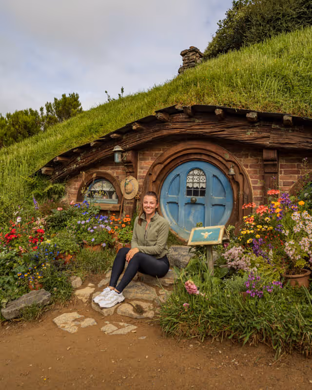 Woman sitting on stone steps in front of a round blue door with a grass-covered roof and colorful flowers around.