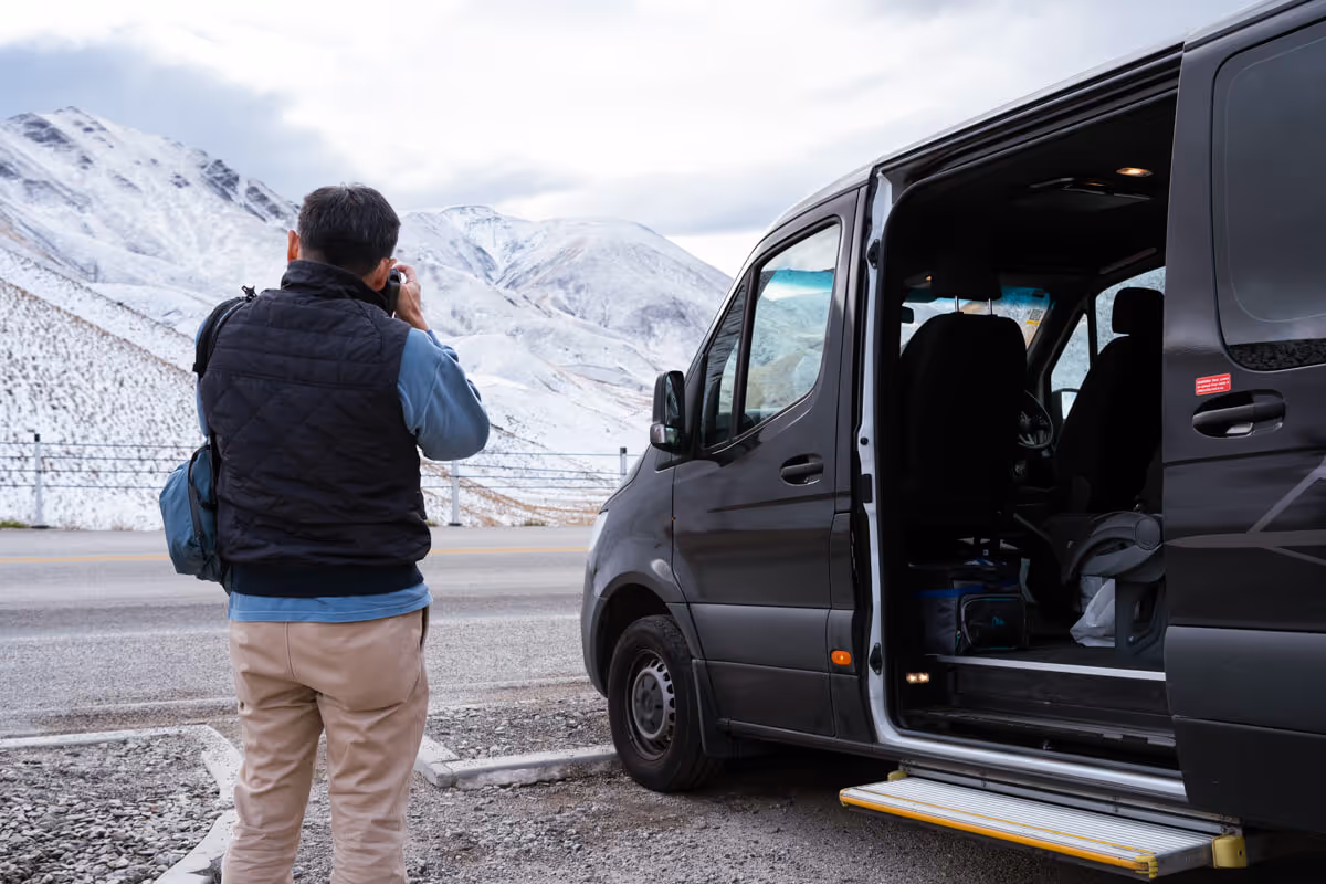 Person taking a photo of snow-covered mountains next to a parked black van with its sliding door open.